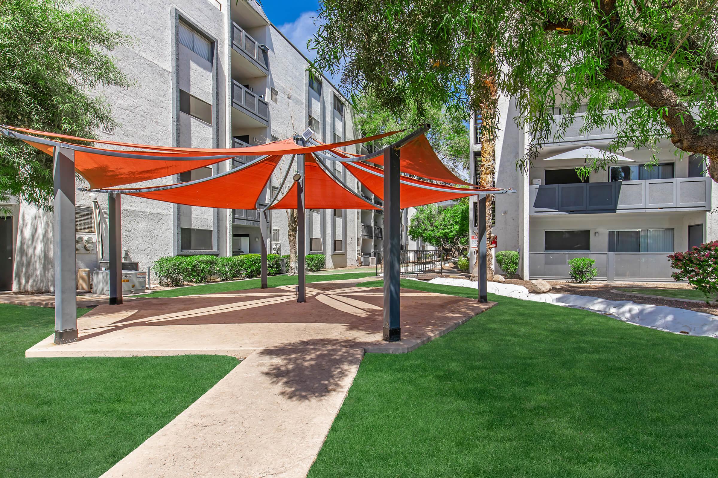 A sunny outdoor area featuring a shaded pavilion with orange canopies. The pavilion is surrounded by lush green grass and small shrubs, with residential buildings in the background. Clear blue skies create a bright, inviting atmosphere.