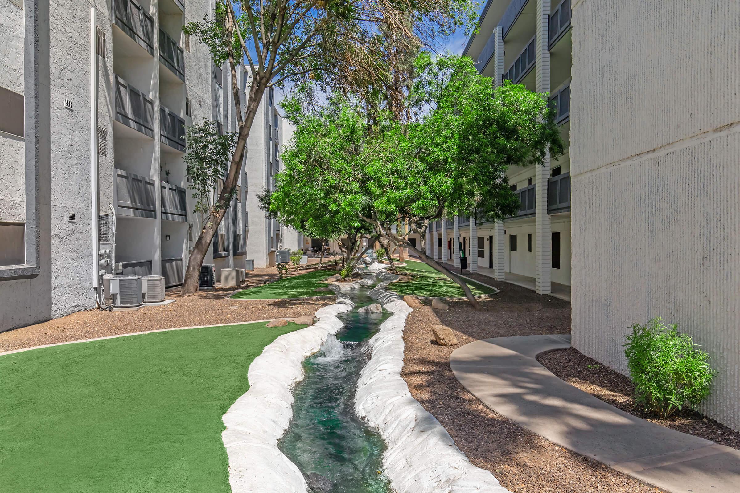 Lush green landscape featuring a small stream winding through a courtyard surrounded by residential apartment buildings. The area is well-maintained with grass and trees, creating a serene outdoor space for residents to enjoy. Bright blue sky adds to the inviting atmosphere.