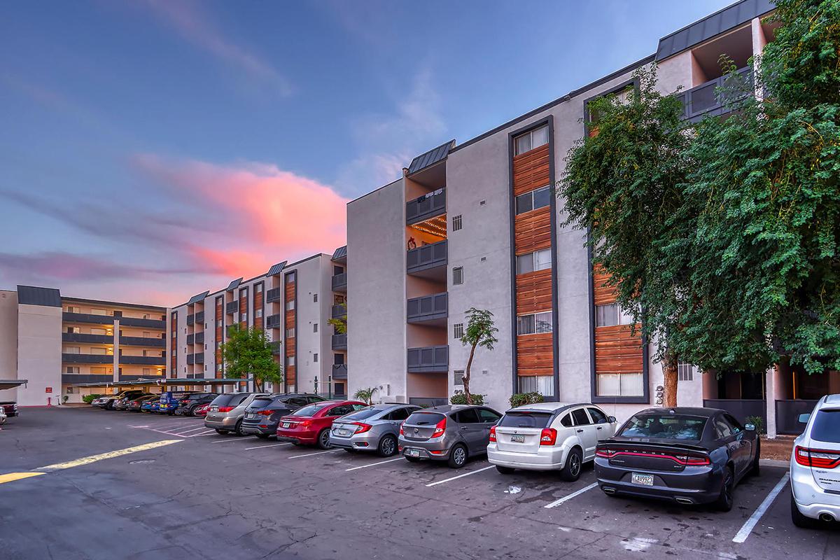 A residential apartment community featuring several multi-story buildings, surrounded by a parking lot with various parked cars. The sky is painted in shades of pink and blue during sunset, creating a vibrant backdrop.