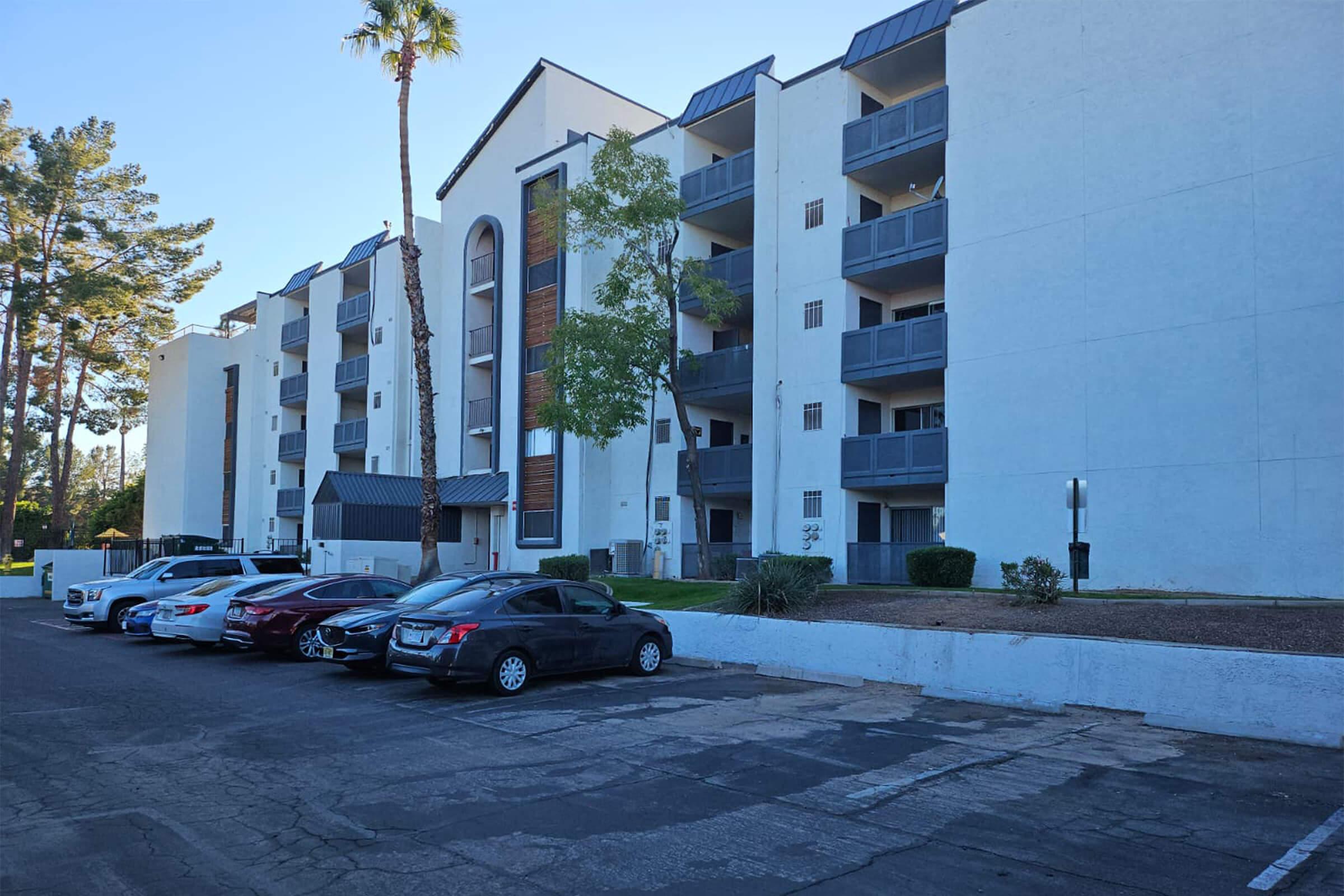 A multi-story residential building with a white facade and balconies on each floor. A row of parked cars in front, with palm trees nearby, under a clear blue sky. The setting appears to be in a suburban area.