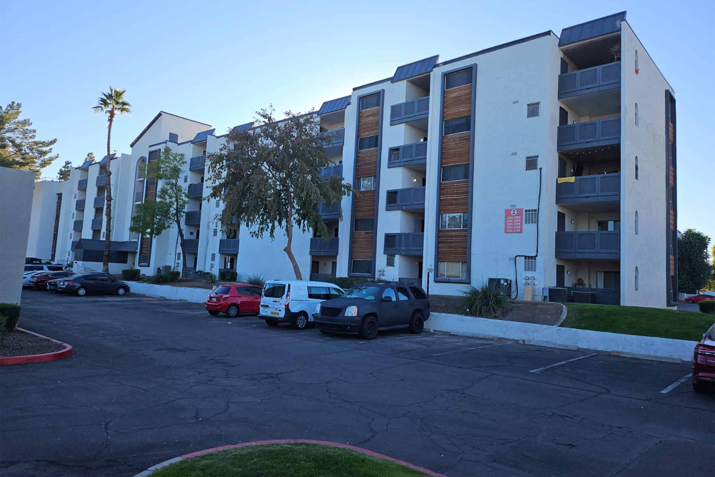 A multi-story apartment building with a mix of balconies and large windows, situated in a parking lot. Several parked cars are visible, including red vehicles and a white van. The surrounding area features trees and greenery, contributing to a residential atmosphere.