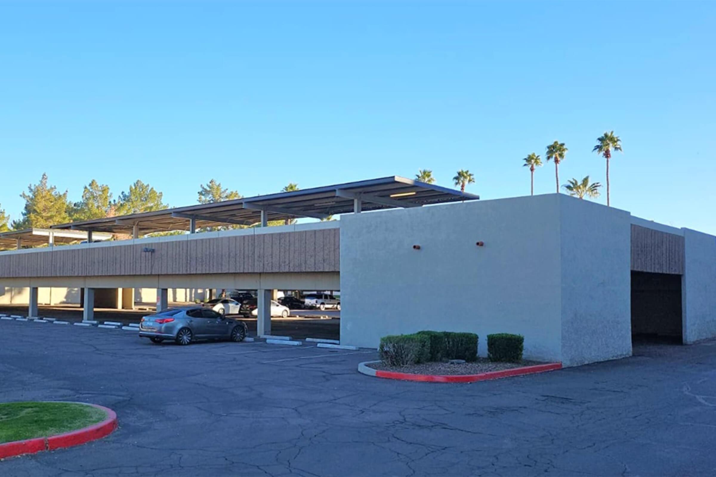 A parking structure with a partially covered area, featuring several parked cars and palm trees in the background against a clear blue sky. The structure has a light-colored exterior with wooden paneling on the upper level. The pavement is cracked, and the landscaping includes low shrubs.