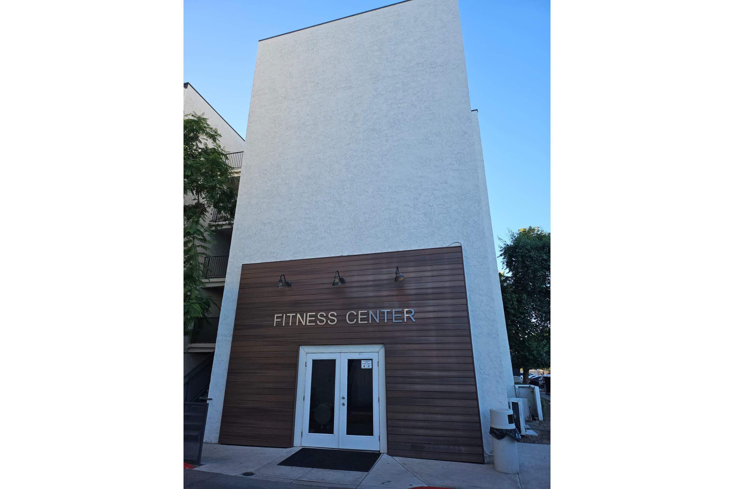 Exterior of a fitness center featuring a simple modern design. The entrance has large double doors beneath a wooden panel with the text "FITNESS CENTER" displayed prominently. Surrounding greenery adds to the inviting atmosphere, and the building rises with a plain white exterior.