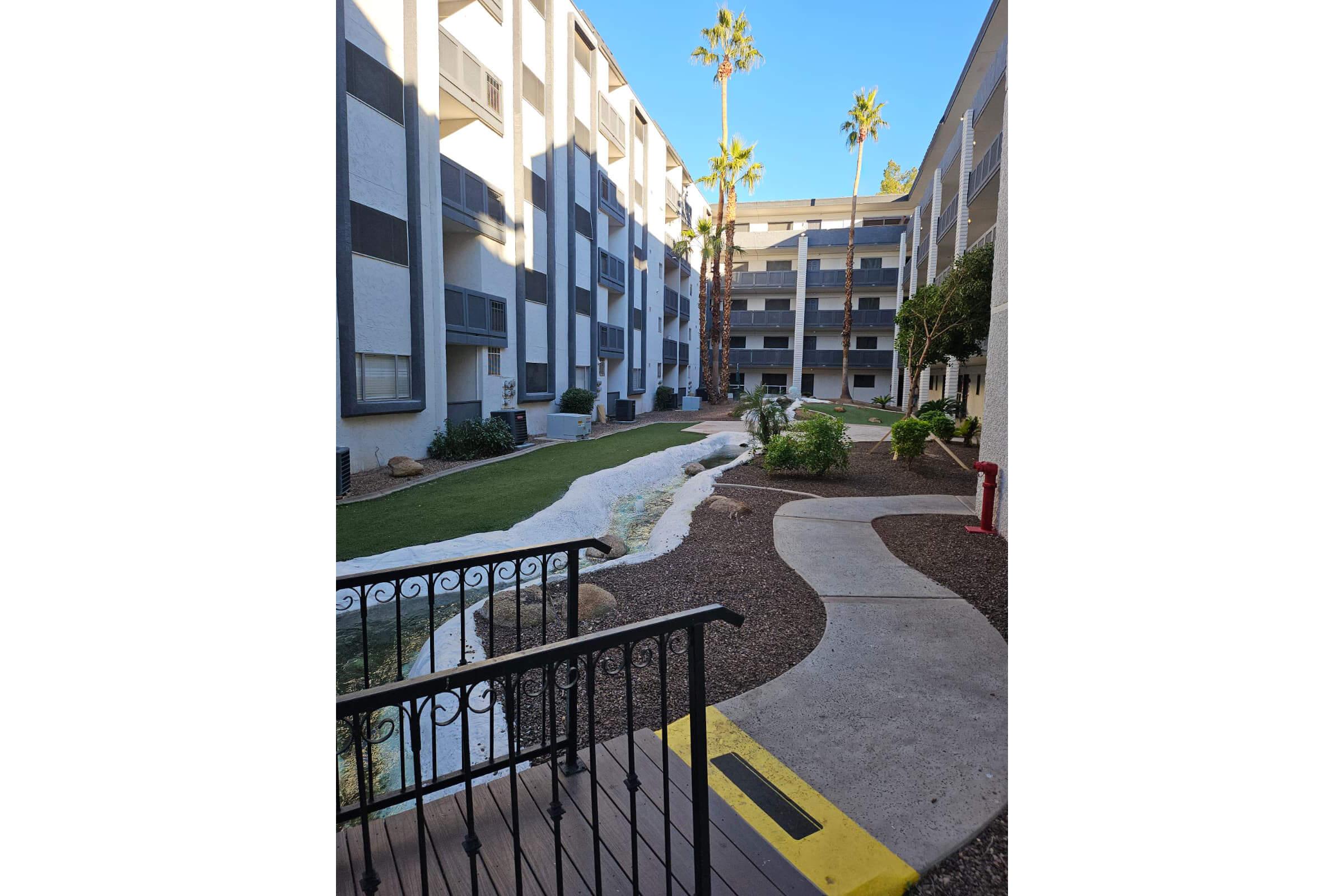View of a courtyard in a multi-story apartment complex, featuring a winding path through landscaped areas with grass, rocks, and palm trees. Natural light brightens the setting, highlighting the buildings on either side, with a small water feature visible in the foreground.
