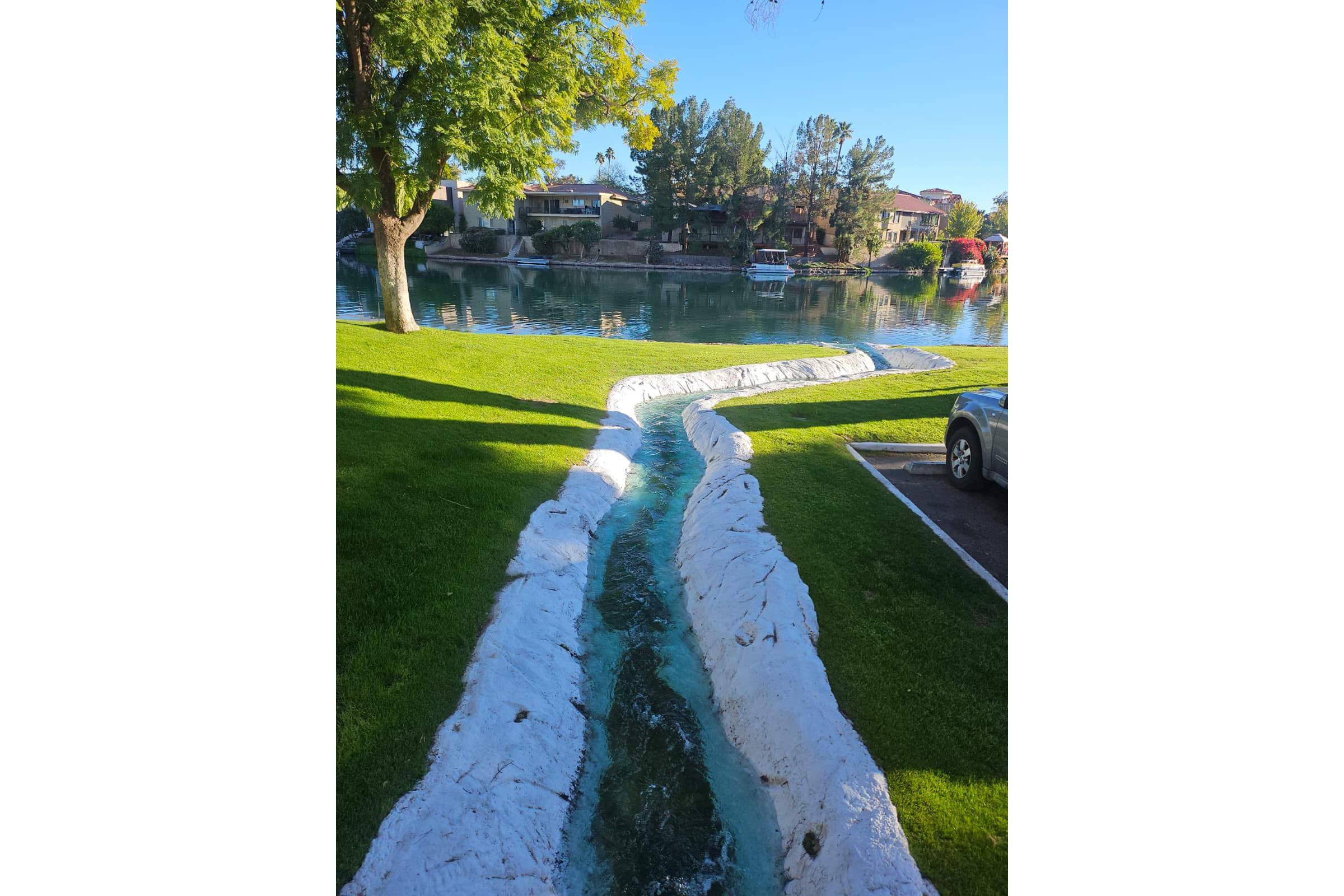 A curved, man-made waterway lined with white stone, running through a grassy area beside a calm lake. The surface of the water reflects the blue sky and surrounding trees, with houses visible in the background. A parked car is partially visible on the right side of the image.