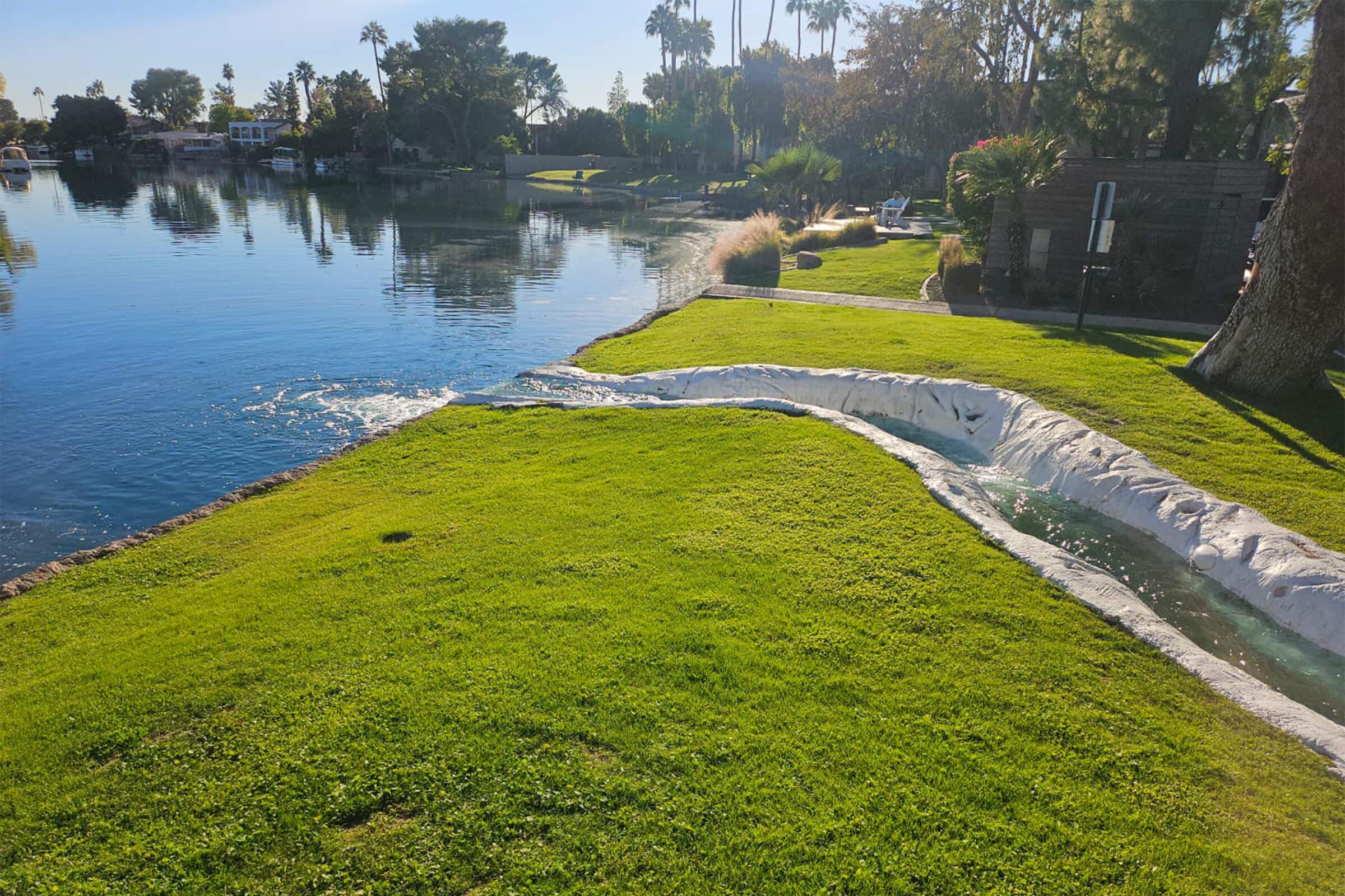 Lush green lawn sloping down to a calm lake, with gentle waves lapping at the shore. Palm trees and landscaping can be seen in the background, reflecting in the water. The scene is serene and picturesque, capturing a sunny day by the water.