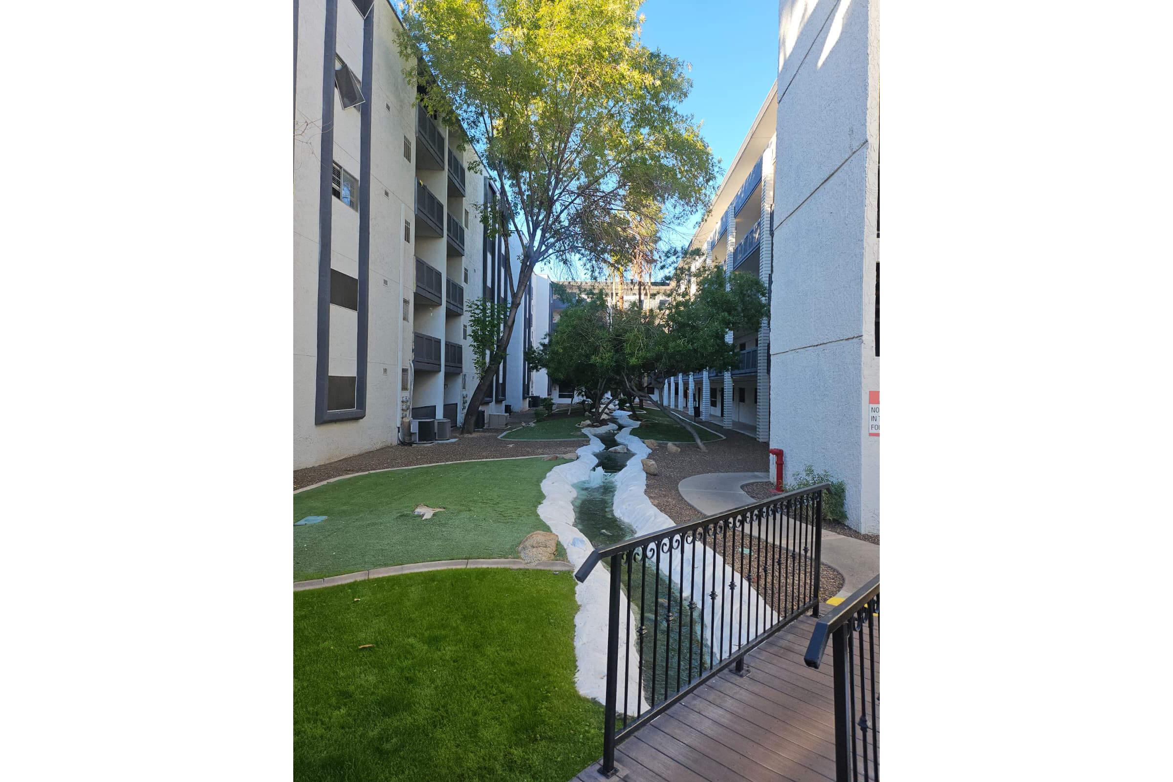 A landscaped courtyard between two apartment buildings, featuring a winding pathway and a small stream. Lush greenery and trees provide shade, and a wooden bridge crosses over the stream. The scene is serene and well-maintained, creating a peaceful outdoor space for residents.