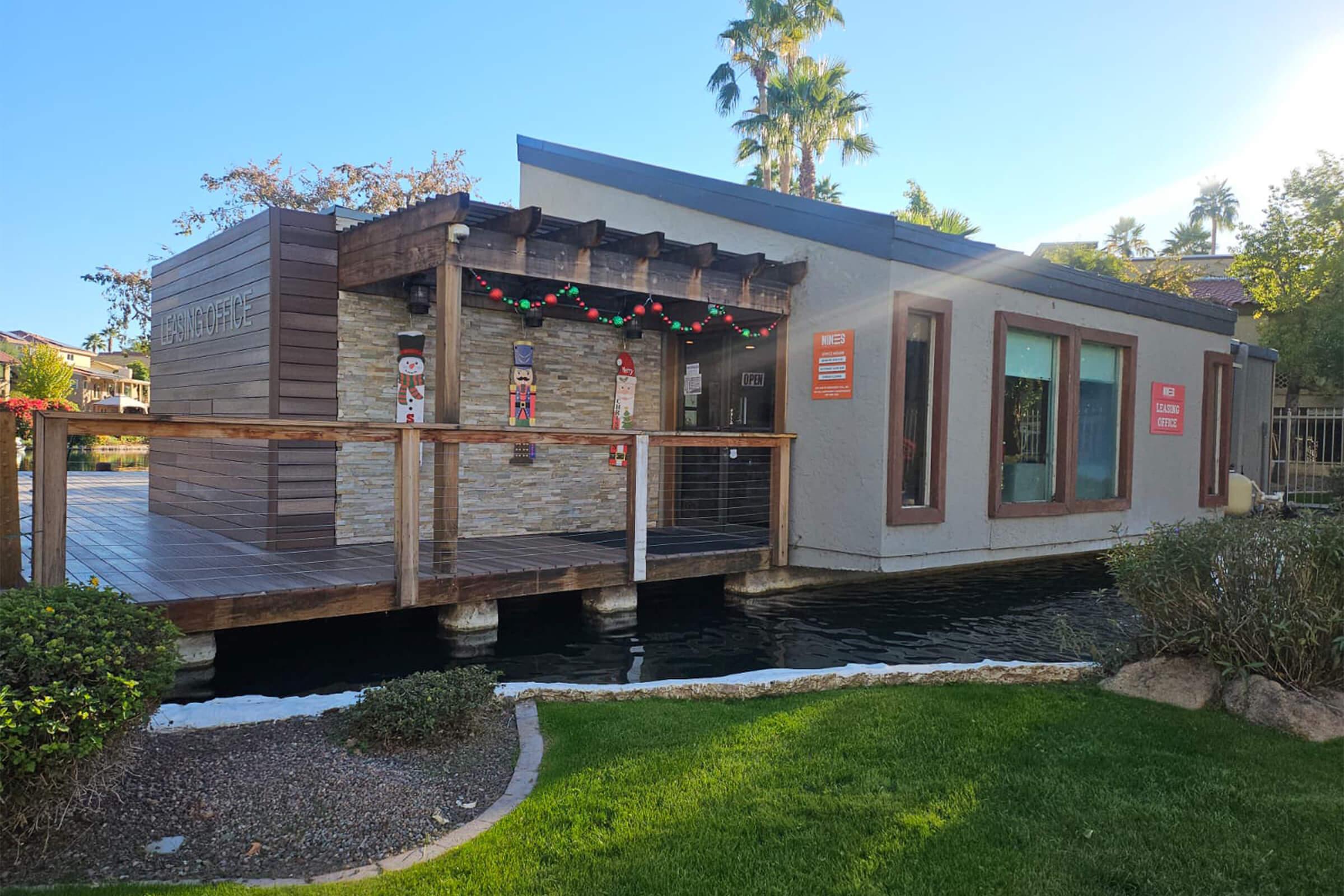 A modern building by the water, decorated with festive holiday lights. The structure has a wooden exterior with stone accents and large windows. Surrounding it are well-maintained plants and a pond. It's a sunny day, showcasing palm trees in the background.