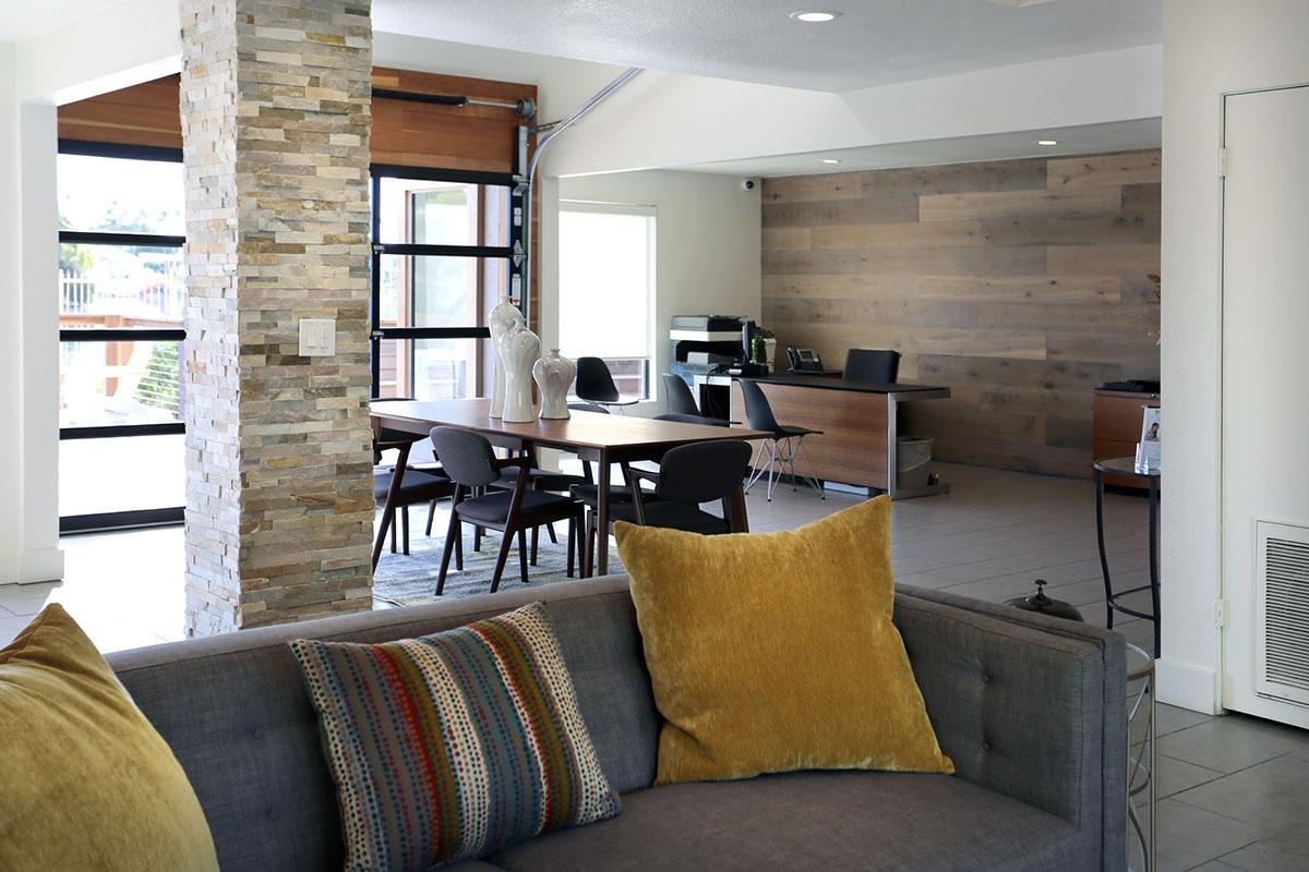 Modern interior of a lounge area featuring a gray sofa with decorative pillows, a wooden dining table with chairs, and an office setup against a wood-paneled wall. Natural light floods the space through large windows.