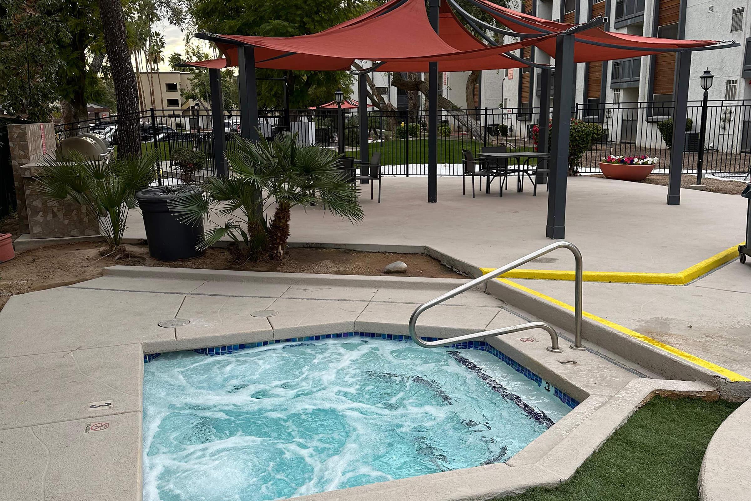 Hot tub with bubbling water surrounded by a concrete patio. In the background, a shaded seating area with red canopies and green grass. Fencing and decorative plants are visible, creating a relaxing outdoor space. The overall setting suggests a residential or recreational area.