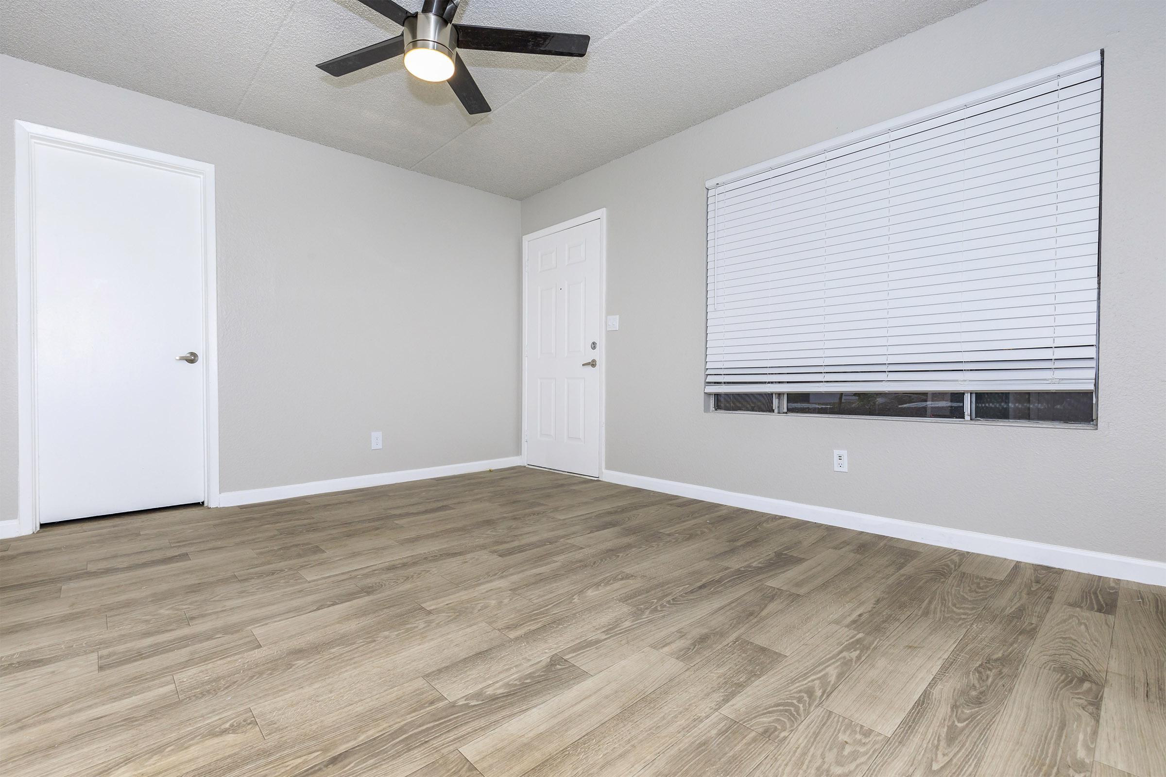 A well-lit, empty room featuring light gray walls, a ceiling fan, and a window with blinds. The flooring is light wood laminate. There is a closed door on the left and a white door leading to another space on the right. The room appears clean and ready for furnishing.