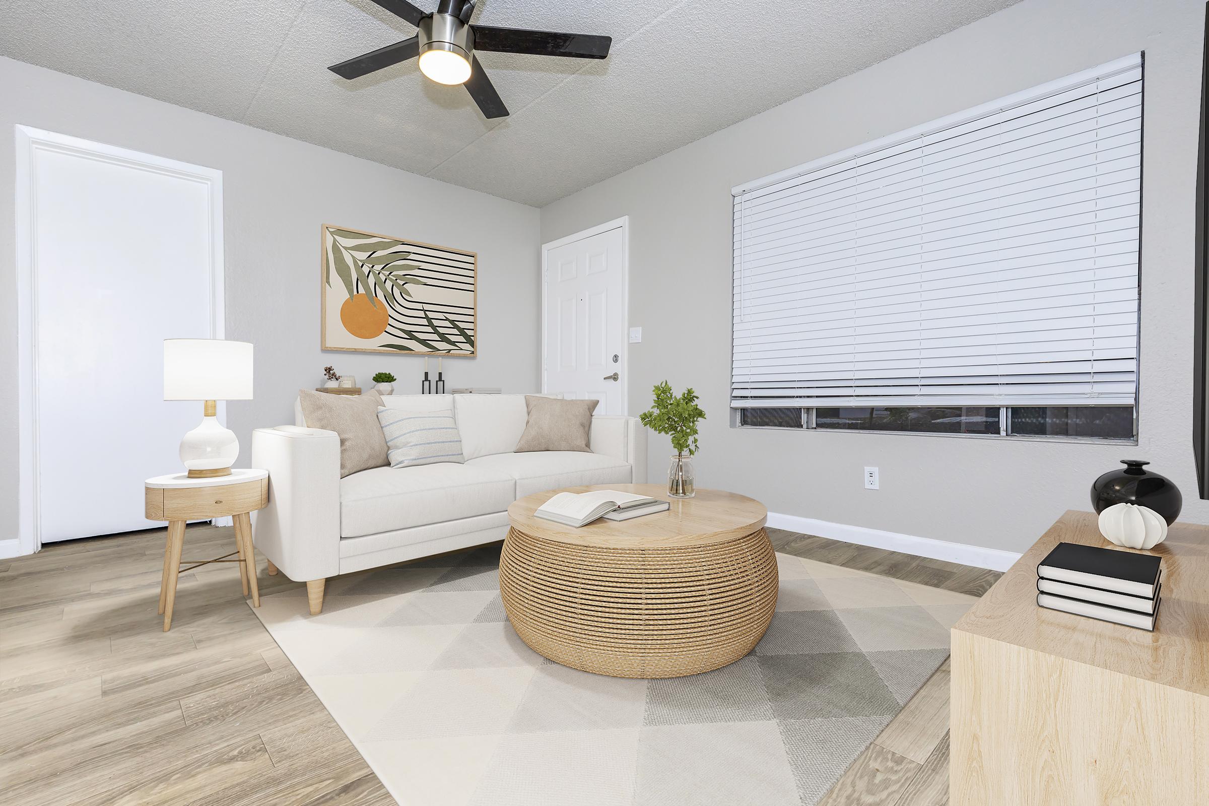 Living room interior featuring a white sofa with pillows, a round woven coffee table, and a light wood side table with a lamp. A potted plant and decorative items are on the table. There is a window with blinds and a piece of abstract art on the wall. The floor is light-colored wood, contributing to a bright, modern atmosphere.