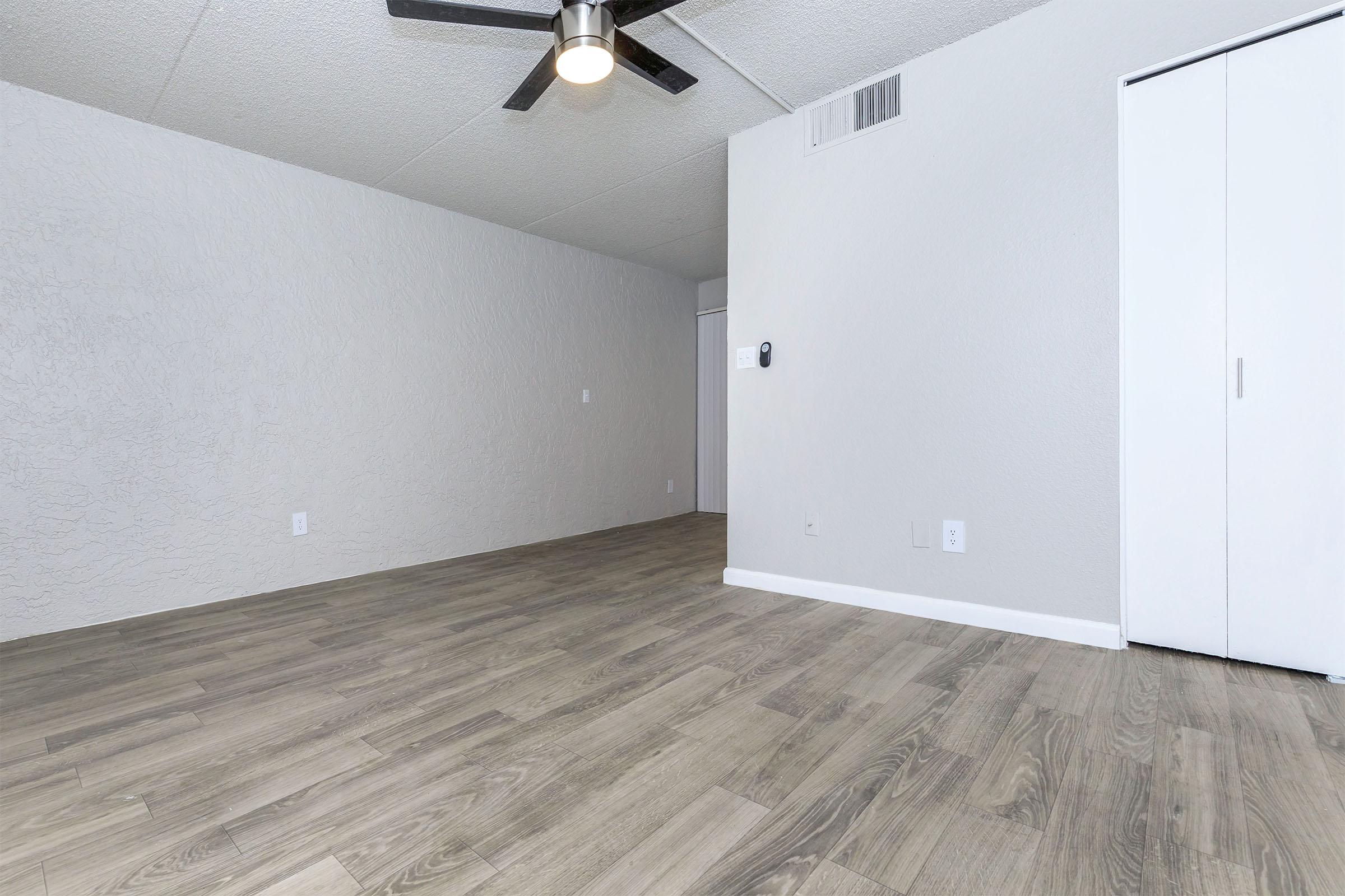 A spacious, empty room with light gray walls and hardwood flooring. A ceiling fan hangs from the ceiling, providing modern aesthetics. The room has a white closet door on the right and a wall-mounted thermostat. The overall ambiance is bright and minimalist, ideal for various interior design options.