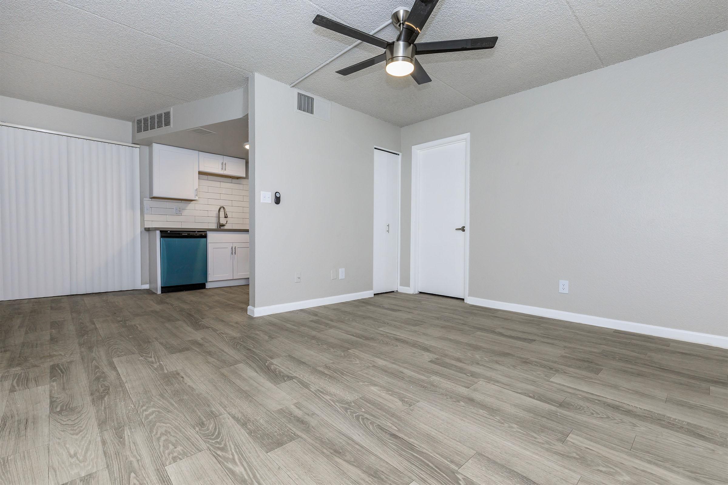 A modern, empty living space featuring light gray walls and a wood-like laminate floor. A ceiling fan is installed, and there is a doorway leading to a kitchen area with white cabinetry and a blue accent. The space is well-lit with natural light and has a clean, minimalist design.