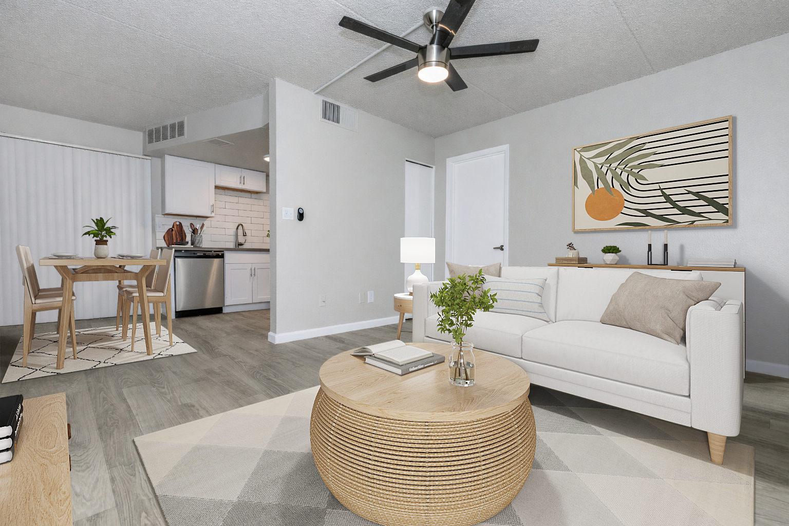 Modern living room with a white sofa and round wooden coffee table. A small plant decorates the table. In the background, there's a dining area with a wooden table and chairs. The kitchen is partially visible, featuring white cabinets and a gray backsplash. A ceiling fan and artwork on the wall complete the cozy ambiance.