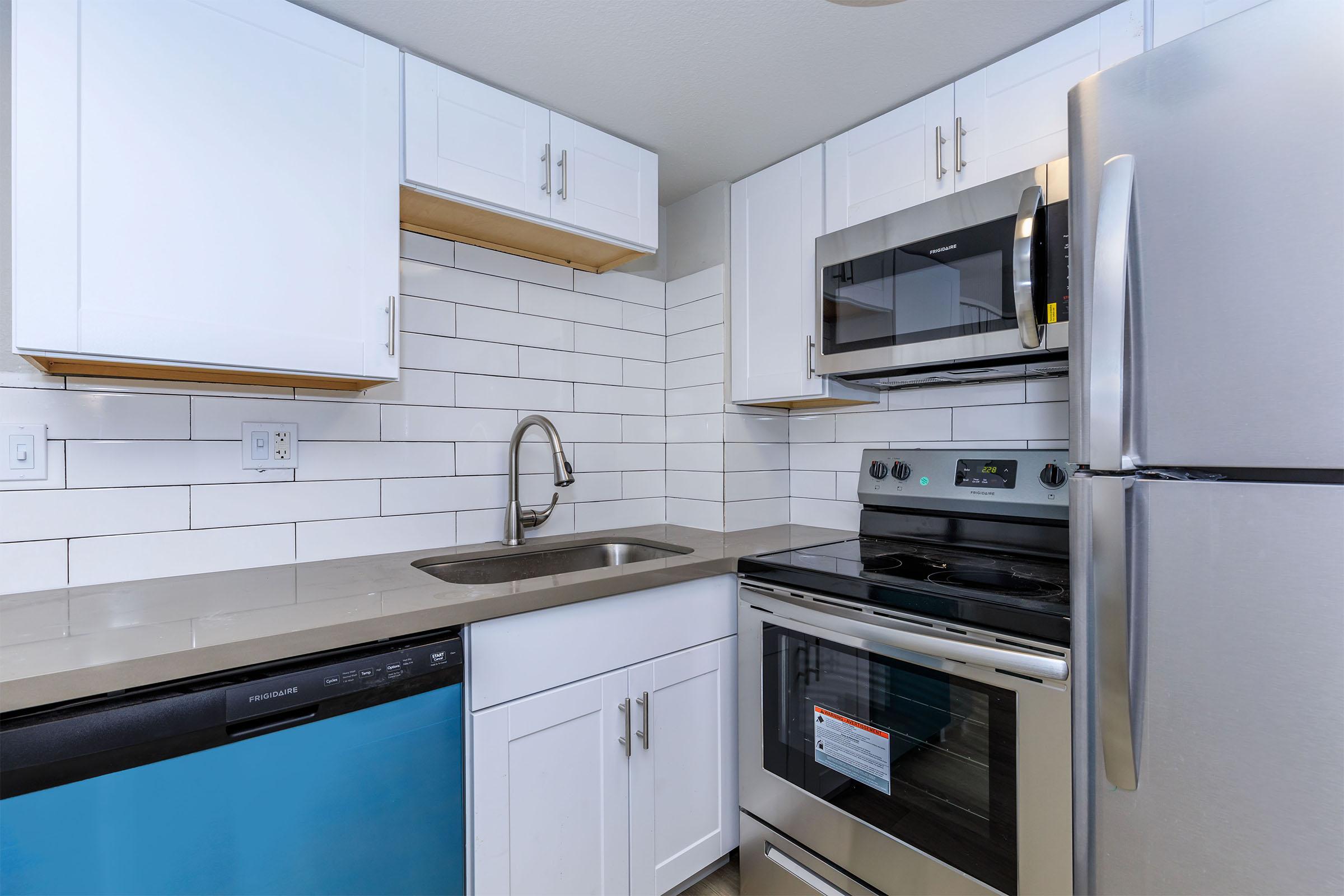 Modern kitchen featuring white cabinetry, stainless steel appliances including a refrigerator, oven, and microwave, and a sleek sink with a faucet. The backsplash consists of white subway tiles, and a dishwasher is visible beneath the countertop, which has a light gray finish.