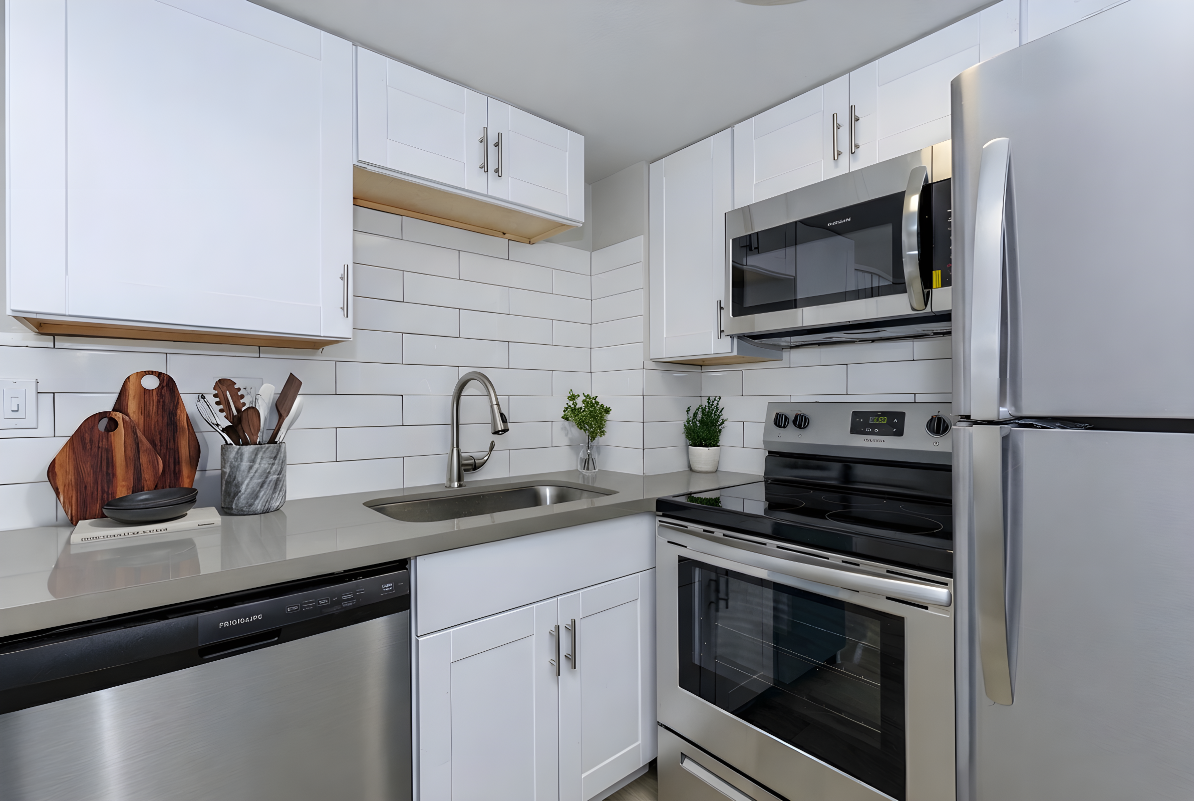 A modern kitchen featuring sleek white cabinetry, stainless steel appliances including a microwave, stove, and refrigerator. It has a gray countertop with a sink and a small plant decor. Kitchen tools and utensils are neatly arranged on the counter, complemented by a stylish cutting board.