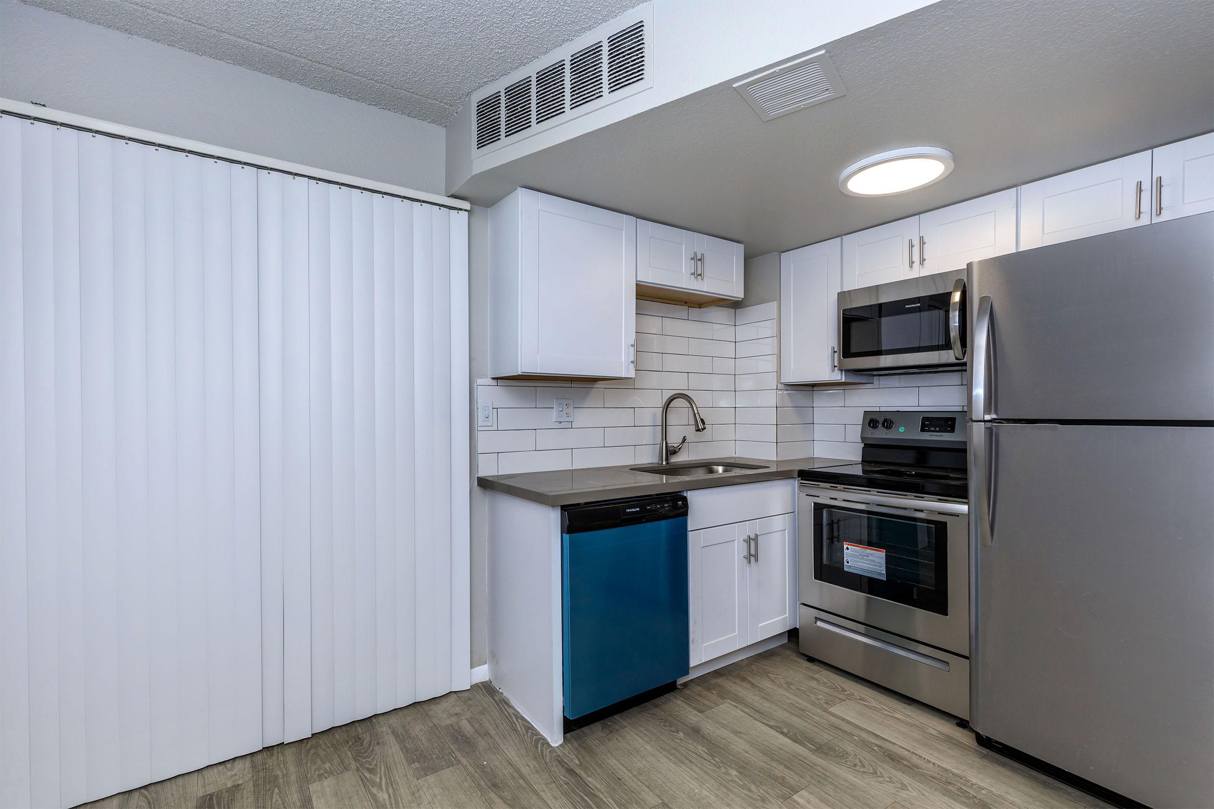 A modern kitchen featuring white cabinetry, stainless steel appliances including a refrigerator, oven, and microwave. The countertop is light gray, with a tiled backsplash. A blue dishwasher adds a pop of color. There's a sliding door with vertical blinds, and the flooring is wood-like laminate.