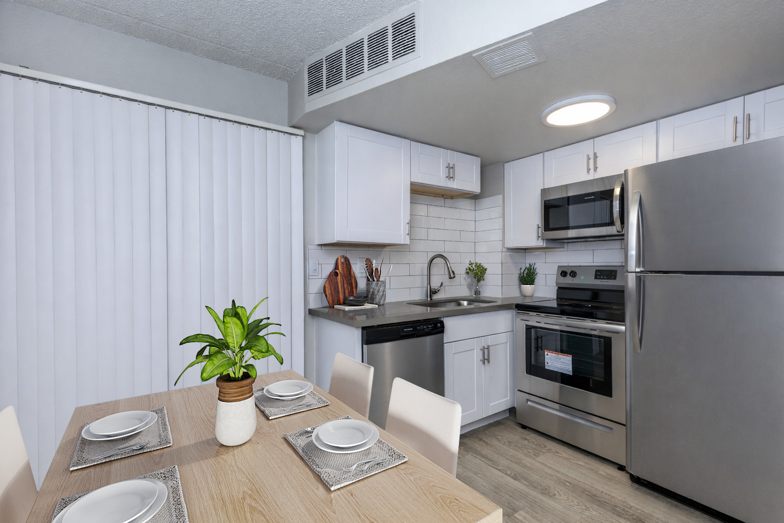 Modern kitchen featuring stainless steel appliances, including a refrigerator, oven, and microwave. White cabinetry and a tiled backsplash complement the space. A dining table with four place settings and a small potted plant add a touch of decor. Vertical blinds are partially drawn on the window.