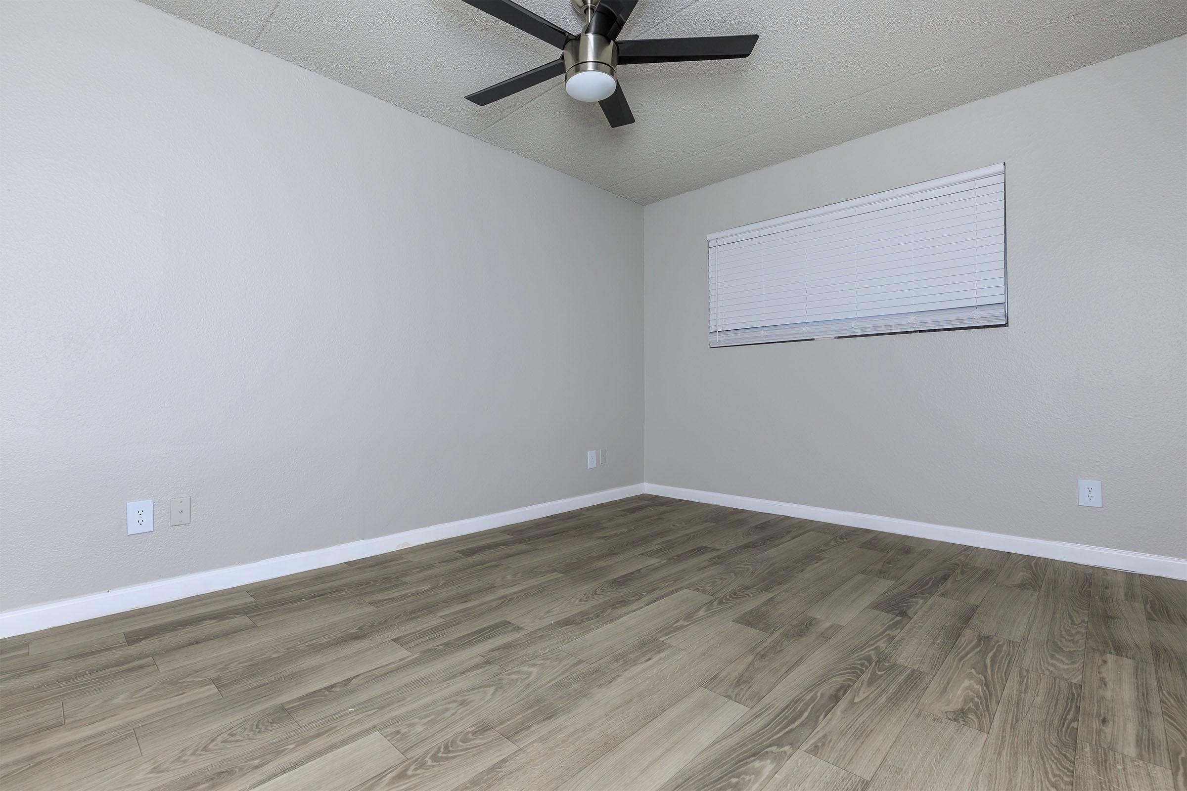 A vacant room with light gray walls and a fan ceiling. The floor features light wooden laminate, and there's a window with white blinds on one wall. The room is well-lit and free of furniture, creating a spacious feel.