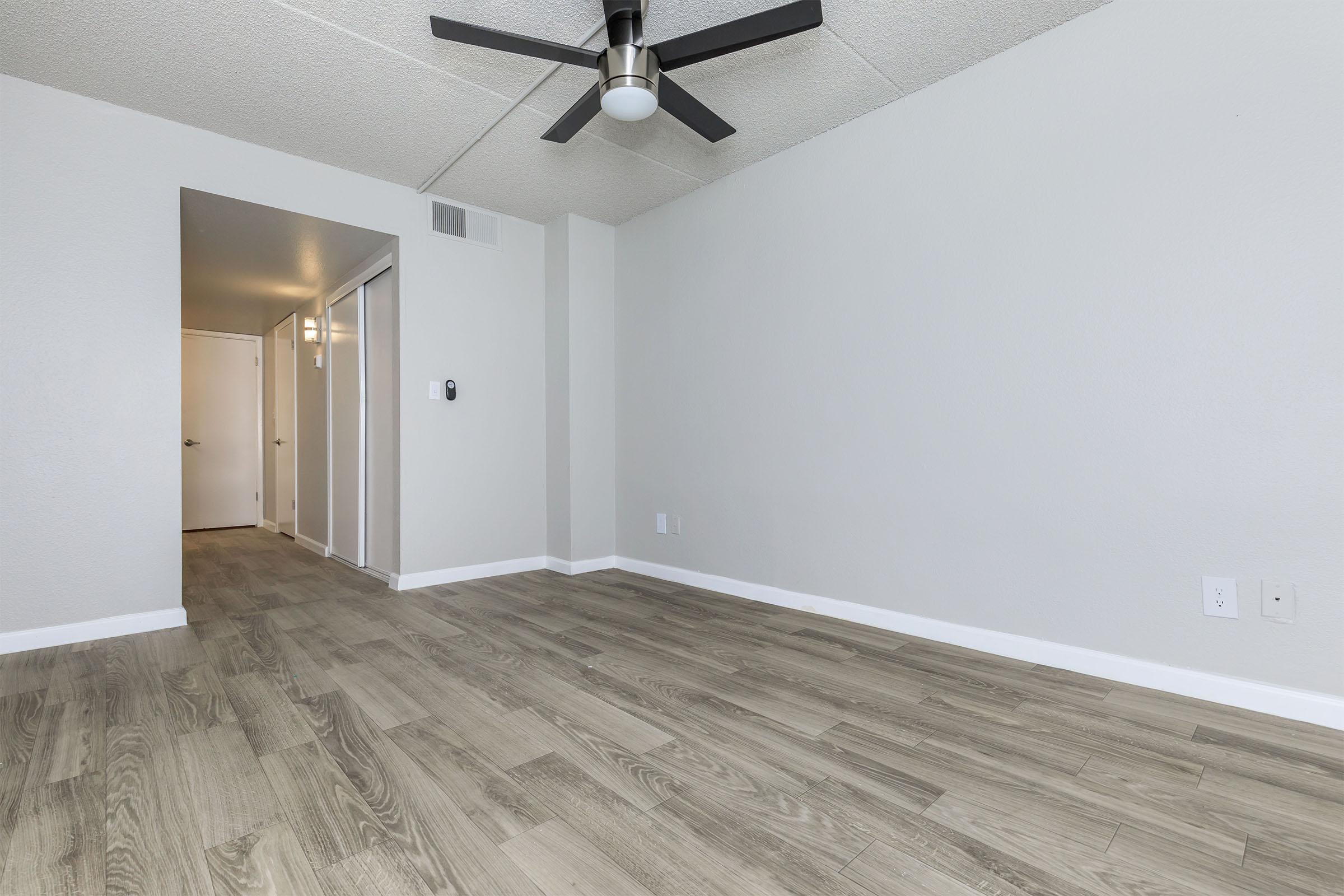 A spacious room with light gray walls and a ceiling fan. The floor is covered with modern wood-style laminate. There is an open doorway leading into a hallway with two closed doors, suggesting a layout for an apartment or home. The overall atmosphere is bright and airy.