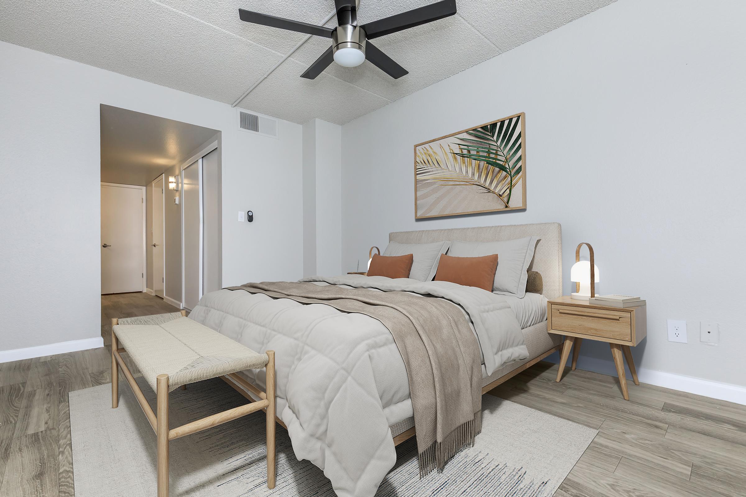 A modern bedroom featuring a beige upholstered bed with brown and orange pillows. There is a light gray textured wall, a ceiling fan, and a wooden bedside table with a lamp. A bench is positioned at the foot of the bed, and a piece of artwork with plant motifs hangs on the wall. The flooring is a light wood tone.