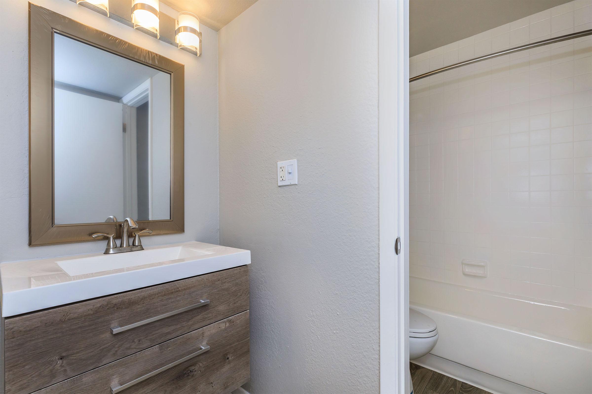 A modern bathroom featuring a light gray wall, a wooden vanity with a white countertop, a mirror with warm lighting above it, and a shower area with a white bathtub and toilet. The design is clean and minimalistic, emphasizing neutral tones and functional space.