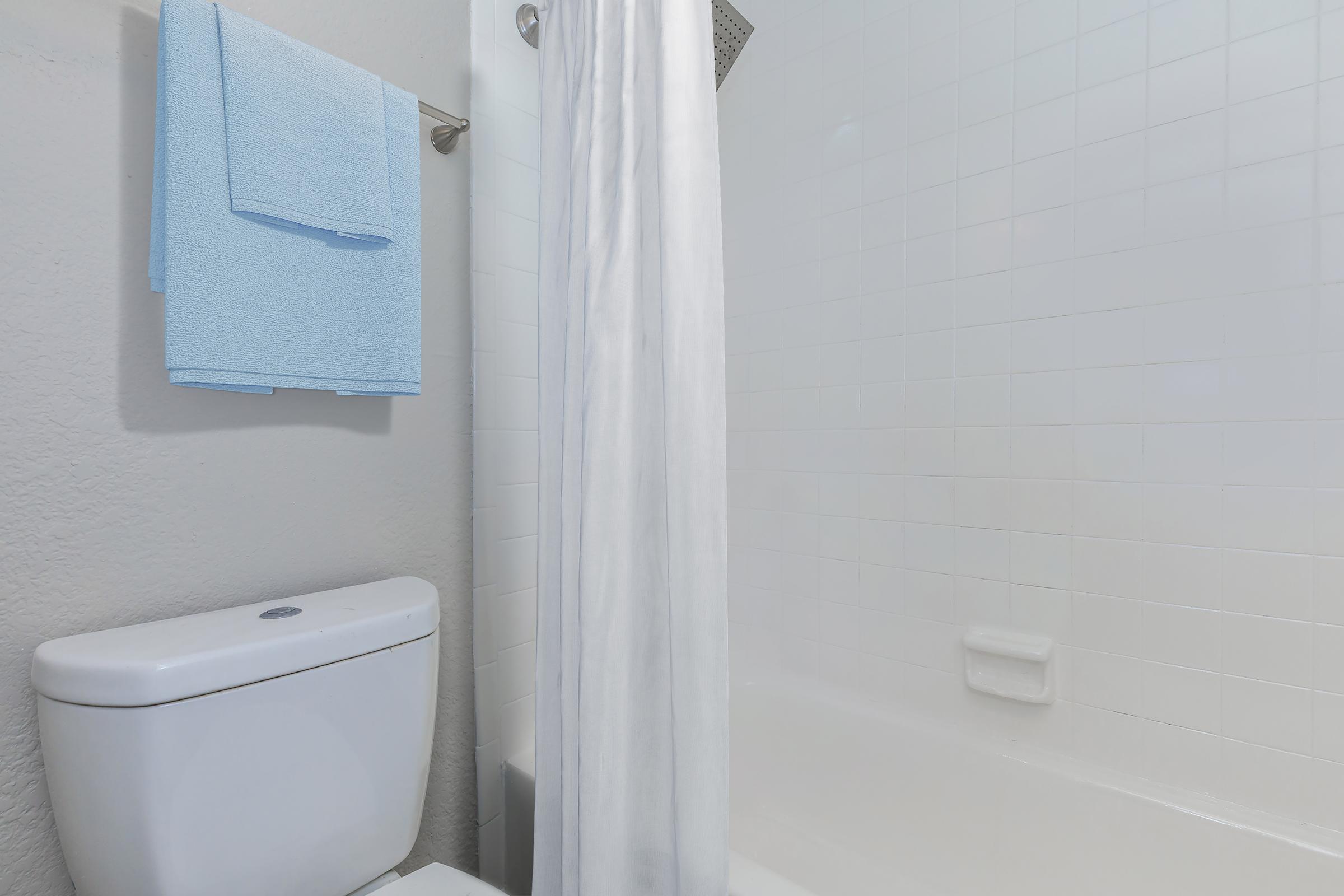 A clean bathroom featuring a white toilet, a bathtub with a shower curtain, and two light blue towels hung on a towel rack. The walls and tiles are white, creating a bright and fresh atmosphere.