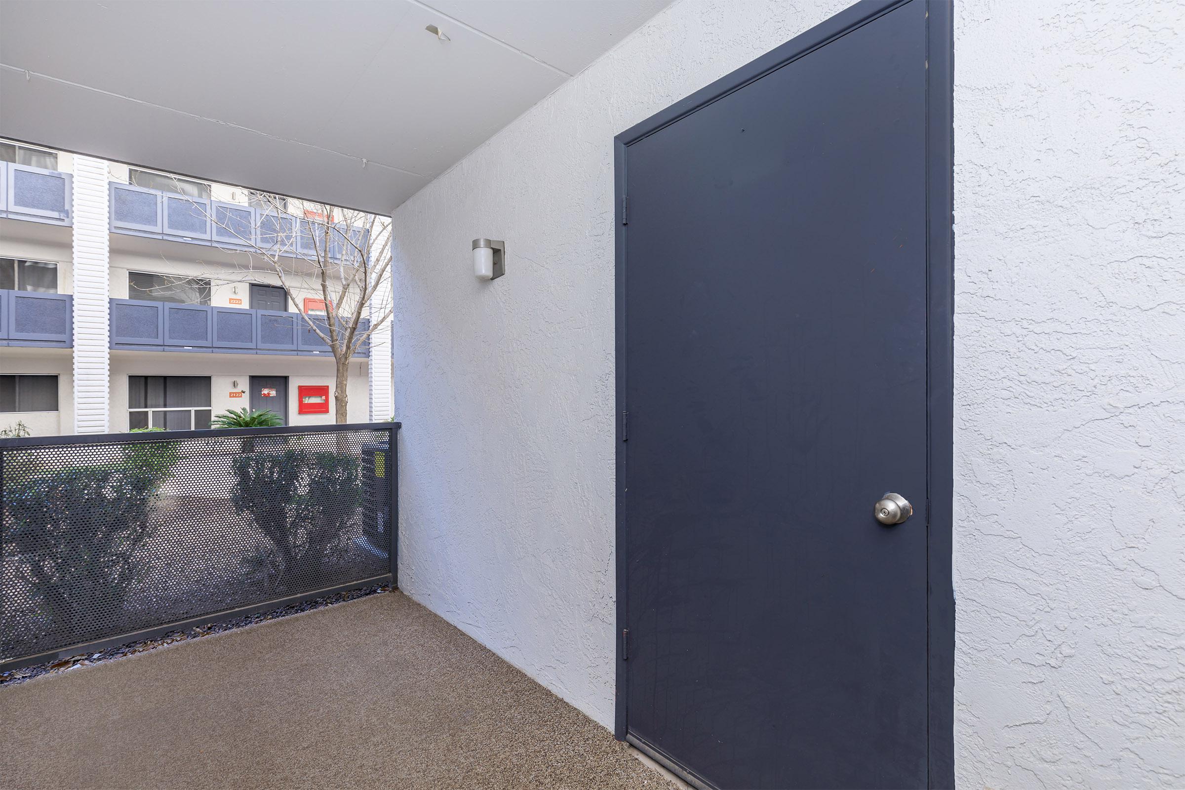 A view of a balcony or porch area featuring a dark door on the right side. The wall is painted white, and there is a light fixture beside the door. In the background, there are apartment windows with a few plants visible, and the floor has a textured surface.