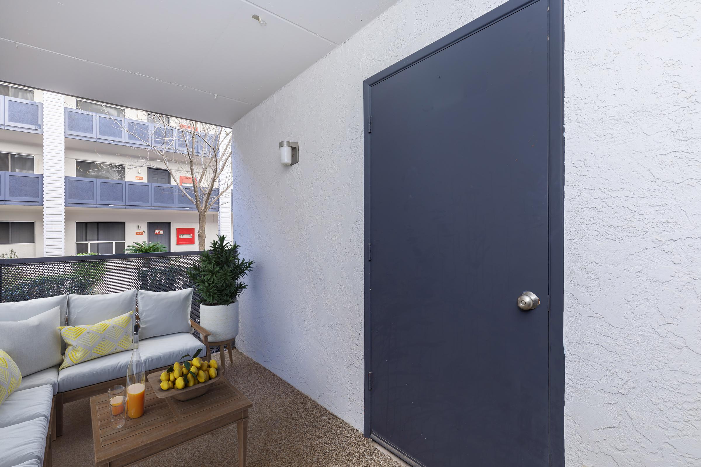 A cozy outdoor patio featuring a small seating area with light blue cushions and a wooden coffee table. On the table, there are two glasses of orange juice and a bowl of yellow bananas. A large potted plant adds greenery, while a closed dark door leads to another space. The background shows a glimpse of an apartment building.