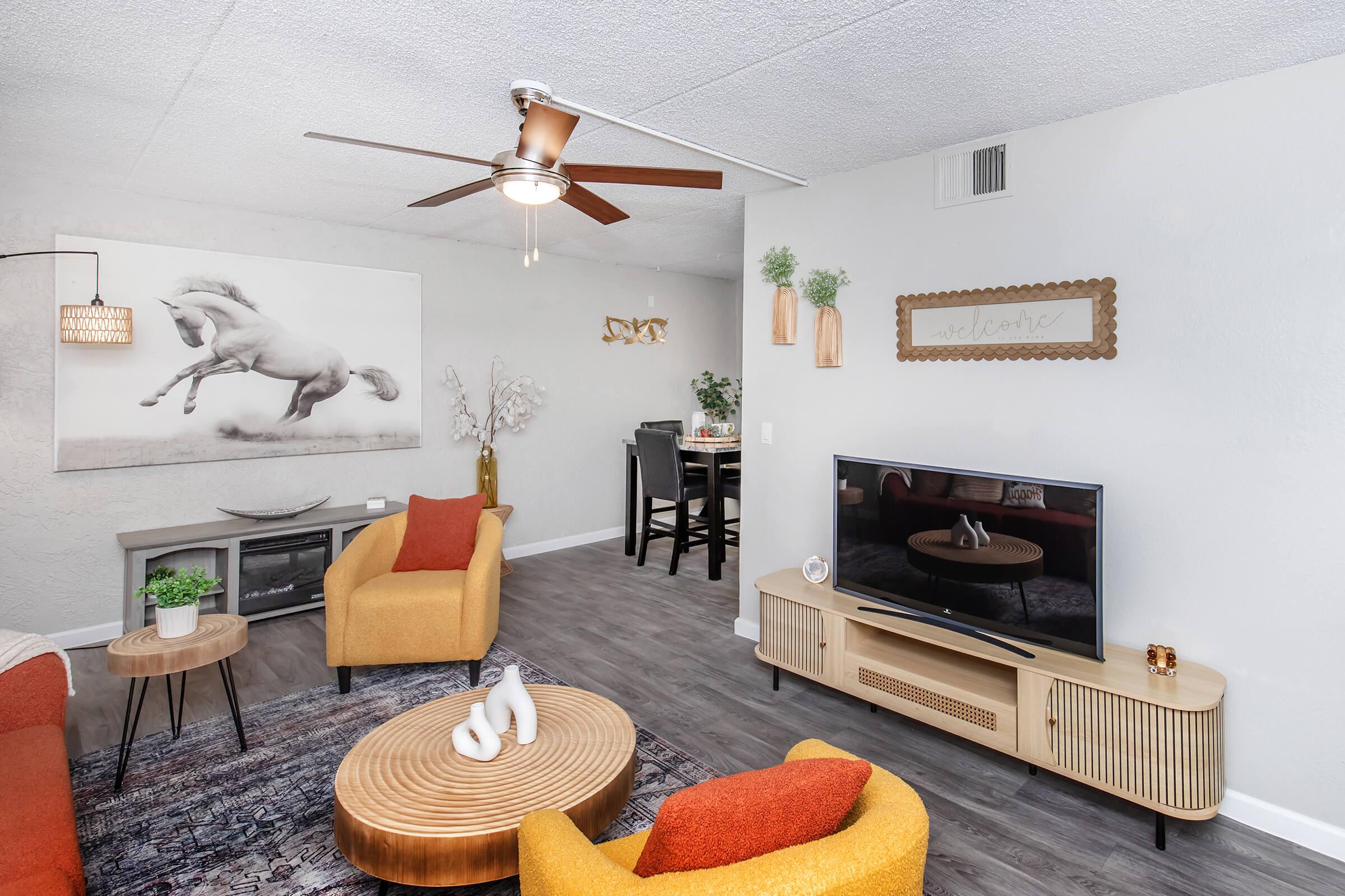A modern living room featuring a ceiling fan, two colorful chairs, a wooden coffee table, and a decorative TV stand. Wall art of a horse is displayed, along with plants and a dining area in the background. The space is bright and inviting with neutral tones and warm accents.