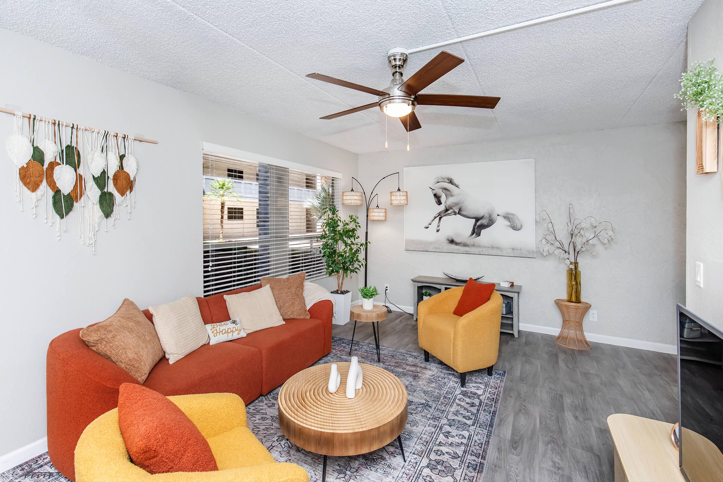 Modern living room featuring a red sofa, two yellow armchairs, and a round wooden coffee table. The decor includes a horse artwork, a ceiling fan, and a wall hanging with leaves. Large windows provide natural light, and the flooring is a light wood finish.