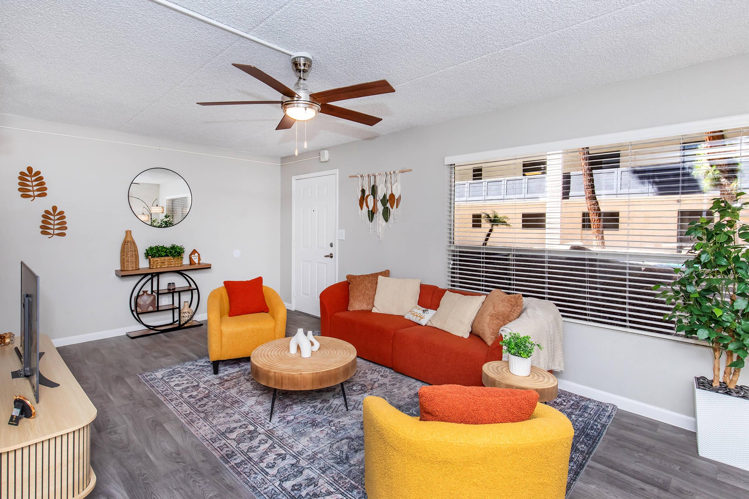 A cozy living room featuring a red and orange sofa with beige throw pillows, two yellow accent chairs, and a round wooden coffee table. A TV stand is on the left, and a mirror is mounted on the wall. Decorative plants and wall hangings add a stylish touch, with natural light coming through the window.