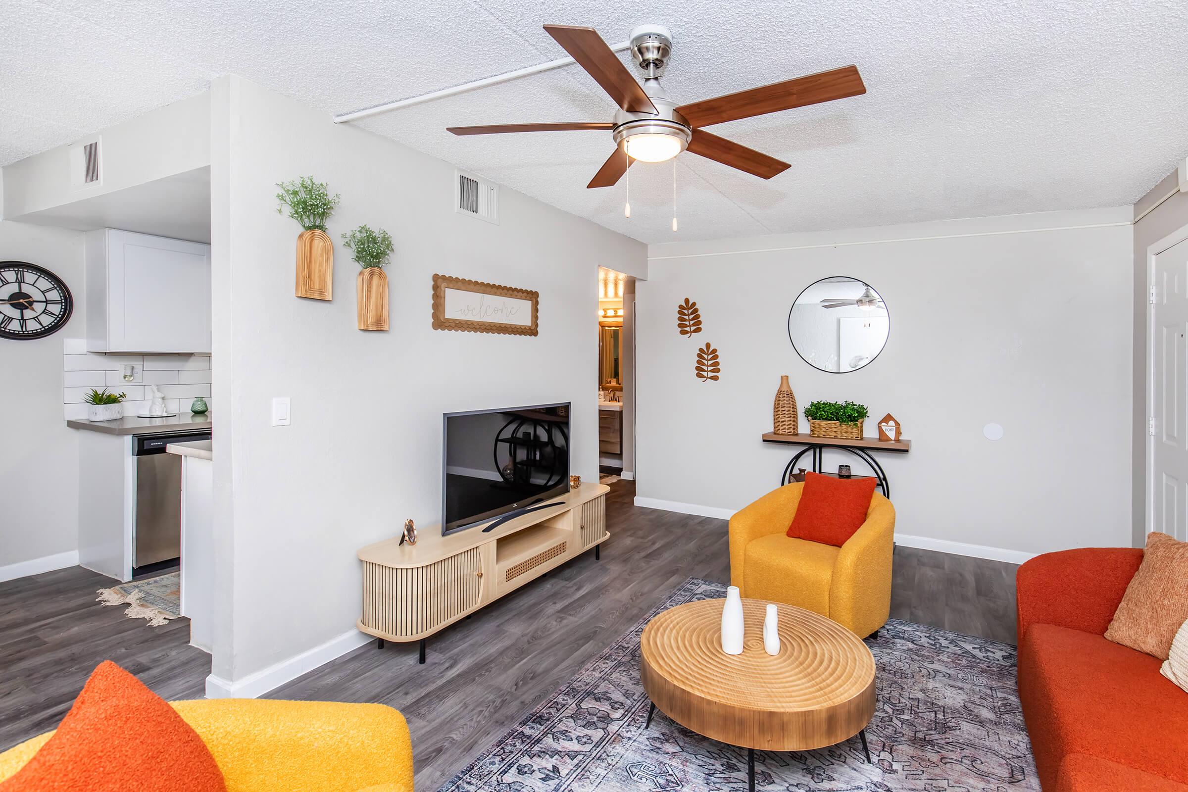 A modern living room featuring a round wooden coffee table, two colorful armchairs in orange and yellow, and a stylish TV stand. The space includes a ceiling fan, decorative wall hangings, and a round mirror. A glimpse of a kitchen area is visible in the background, adding to the home's inviting atmosphere.