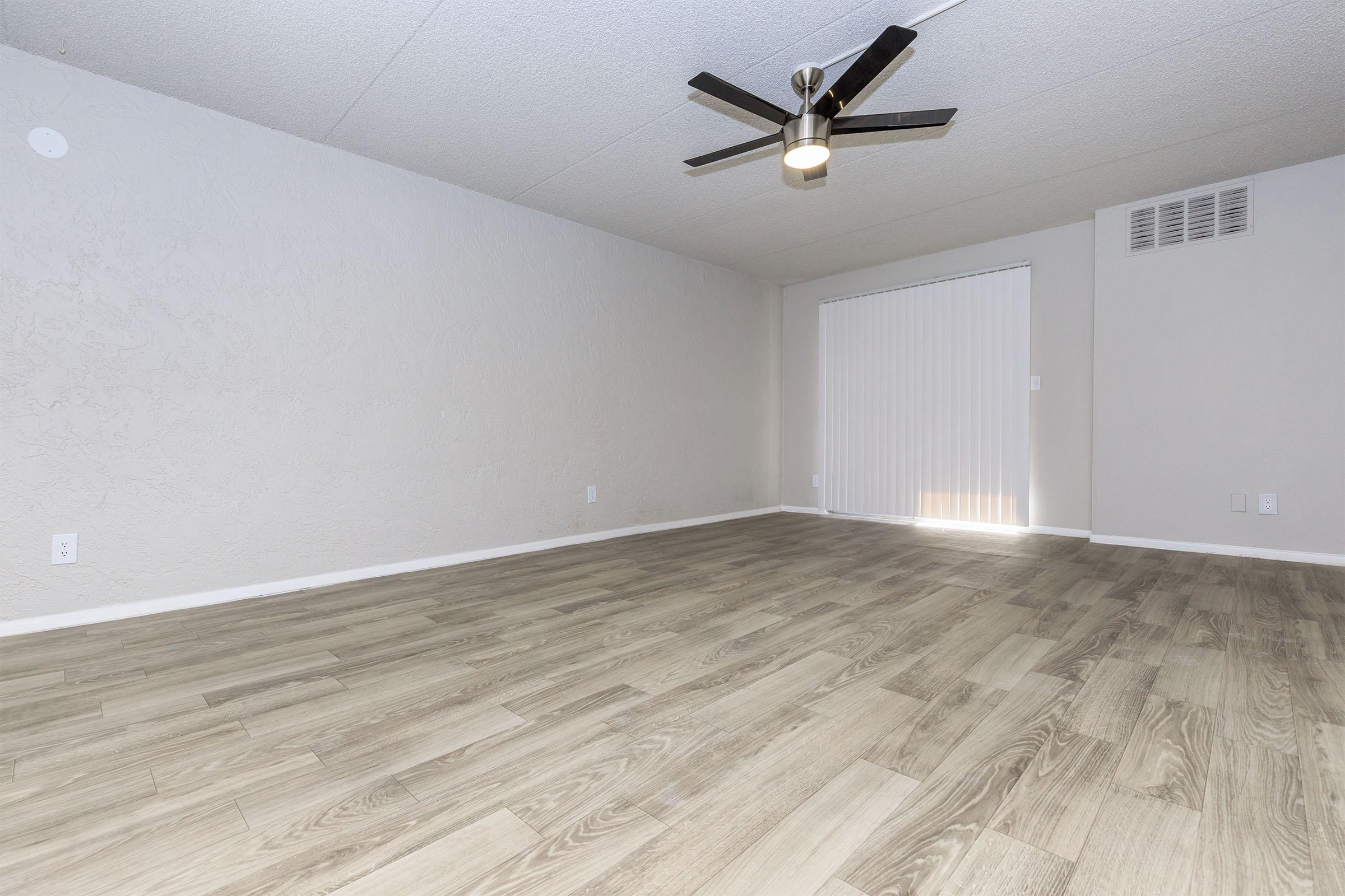 Spacious interior of a room featuring light-colored walls, a wood-like floor, and a ceiling fan. There is a sliding door with vertical blinds allowing natural light to enter from outside. The overall atmosphere is bright and clean, ideal for various furnishings and decor.