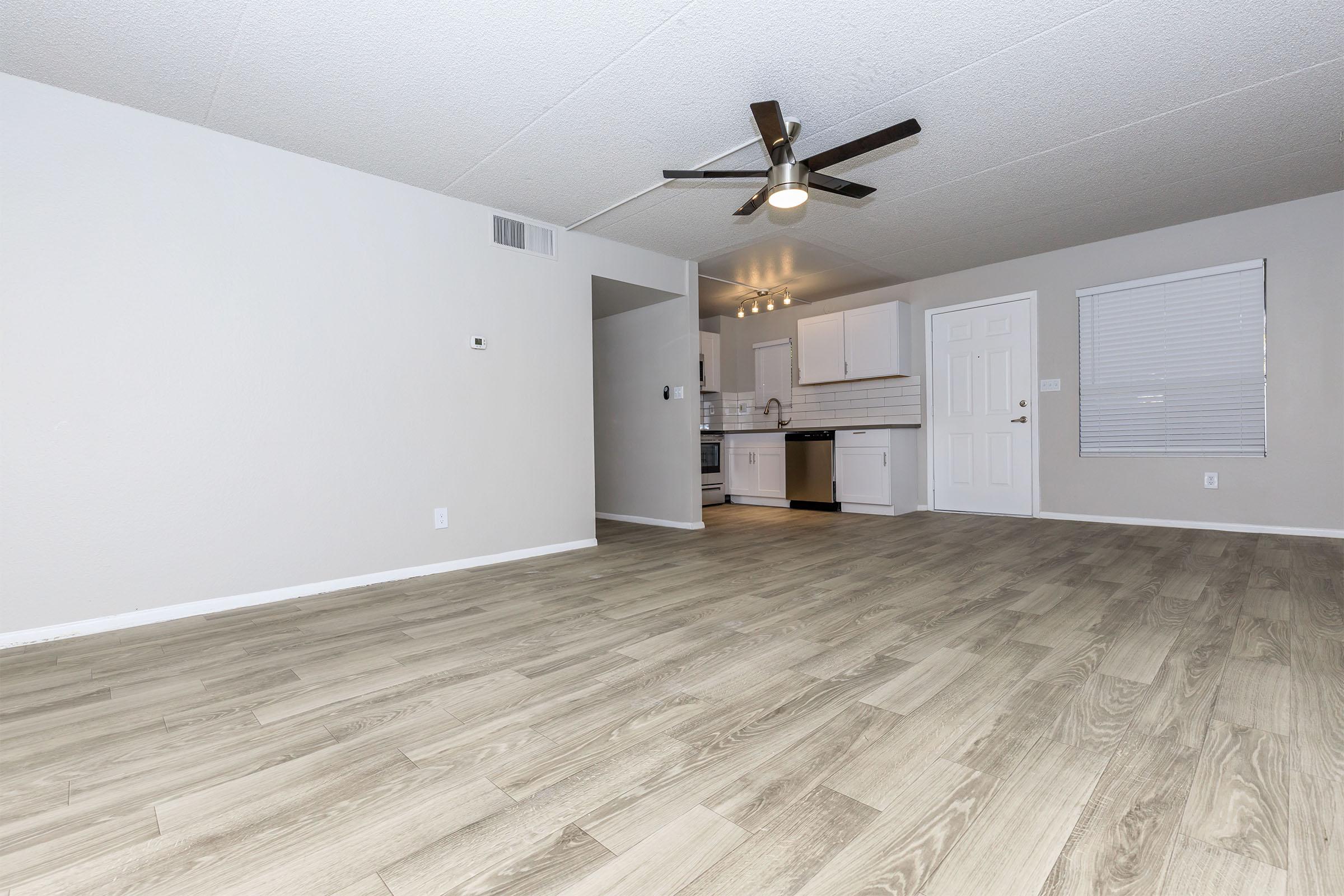 A spacious, modern living area with light-colored, wood-like flooring. A ceiling fan is mounted above, and there's a doorway leading to a kitchen area with white cabinets and appliances. The walls are painted in neutral tones, providing a bright and inviting atmosphere. A small window allows natural light to enter.