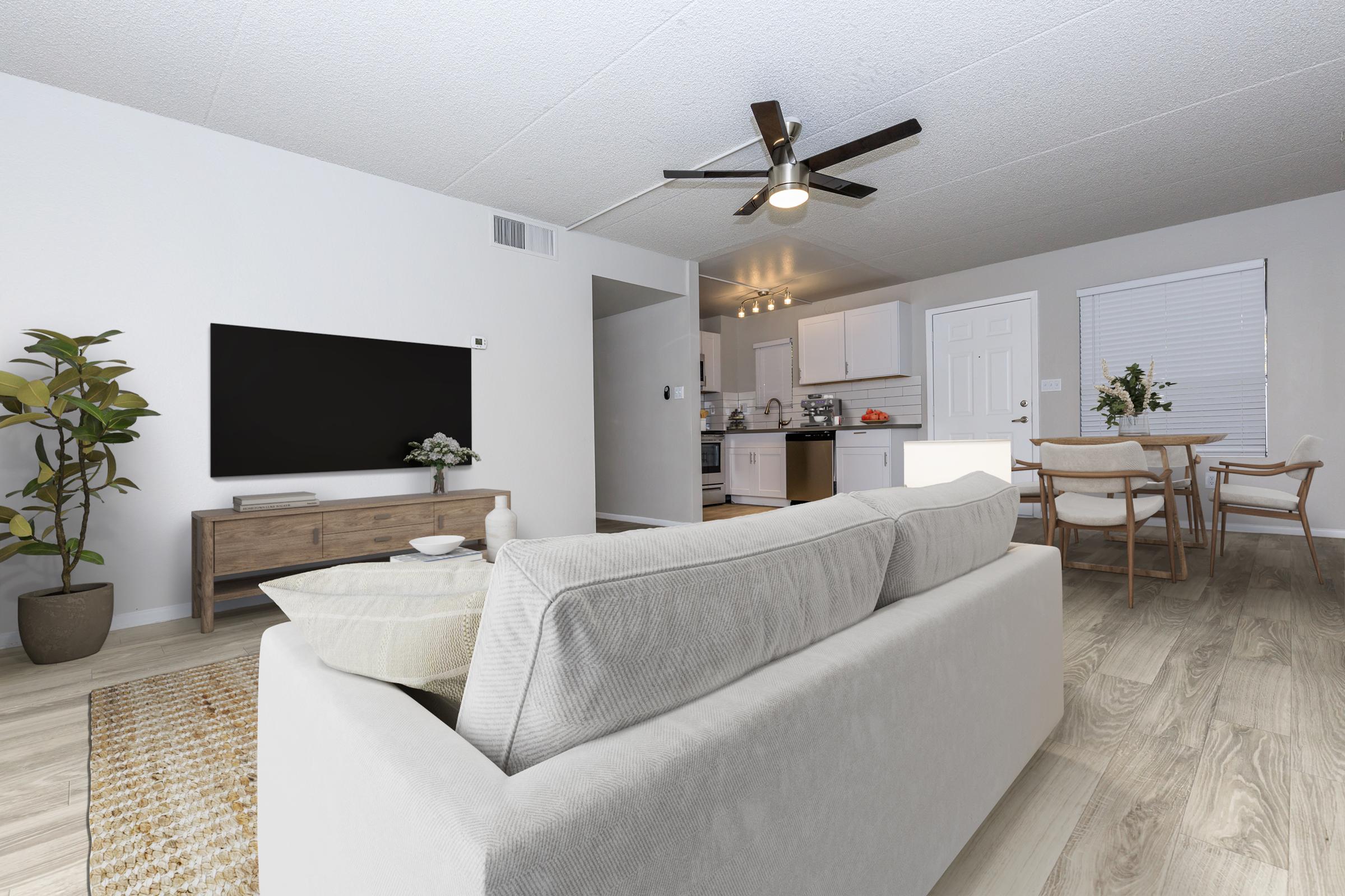 A modern living room featuring a light-colored sofa with a decorative throw pillow, a wooden TV stand with a large flat-screen TV, and a potted plant. The open kitchen is visible in the background, showcasing white cabinets and dining furniture. The space has a bright, contemporary feel with light wood flooring.