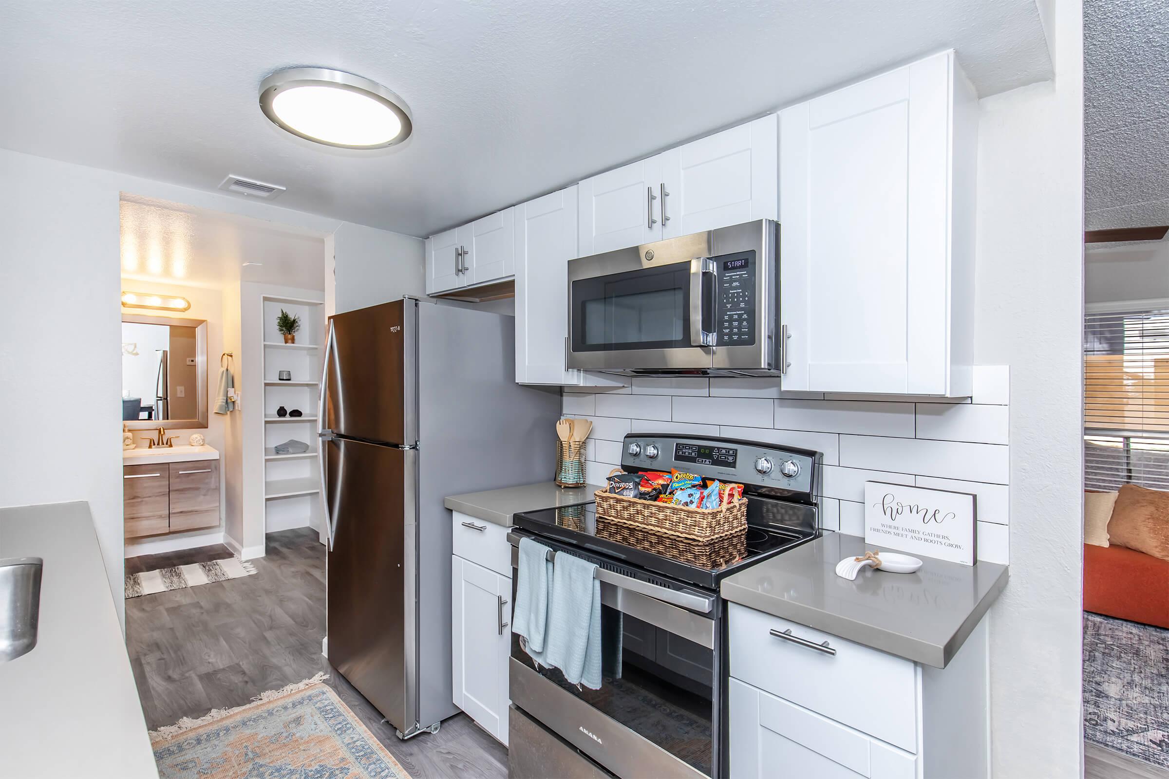 Modern kitchen featuring stainless steel appliances, including a stove, microwave, and refrigerator. The white cabinets complement the gray countertops, and a basket sits on the stove. The room is well-lit with a ceiling light, and a glimpse of a bathroom and living area can be seen in the background.