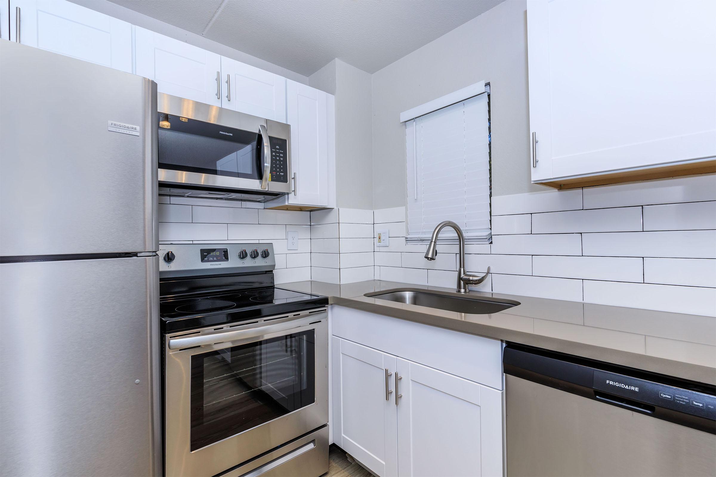 Modern kitchen with stainless steel appliances including a refrigerator, microwave, and oven. The countertop is a light gray color, and there are white cabinets above and below. A stainless steel sink is installed beneath a window with white blinds, and the wall features white subway tiles.