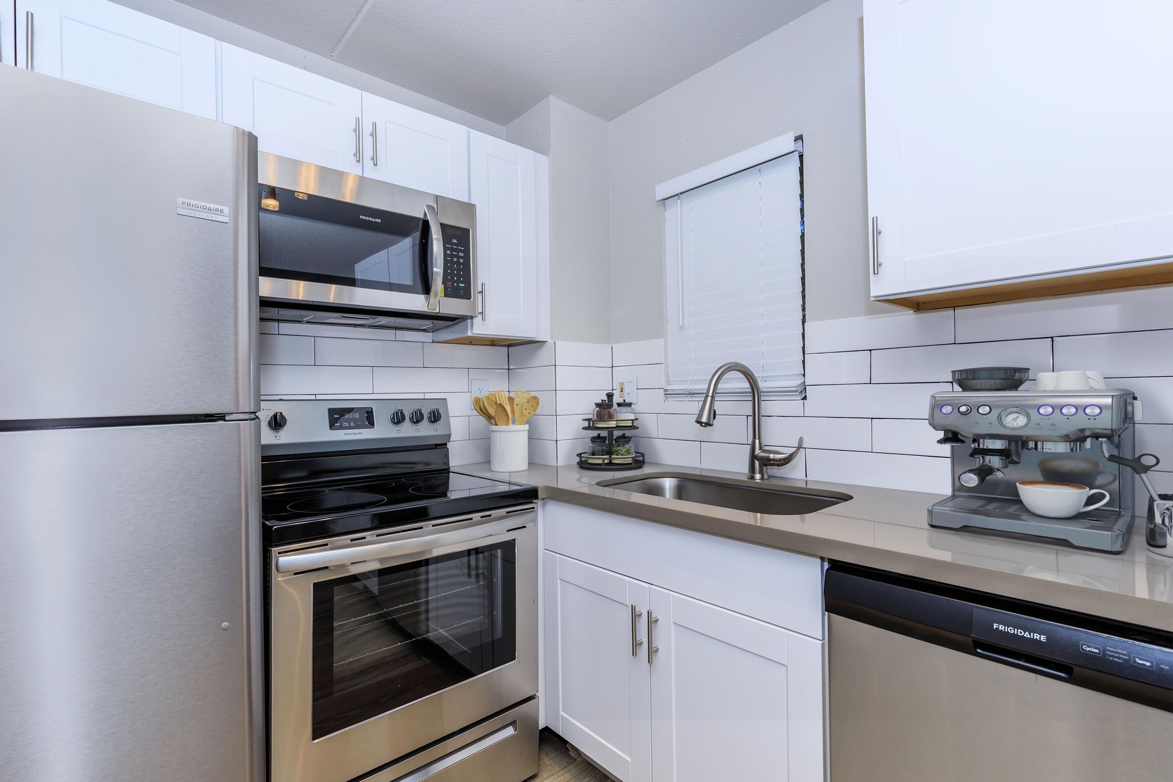 Modern kitchen featuring stainless steel appliances, including a refrigerator, microwave, oven, and dishwasher. The white cabinets and gray countertops create a sleek look. A coffee machine is on the counter beside a sink, and utensils are displayed in a holder. Modern tile backsplash completes the design.