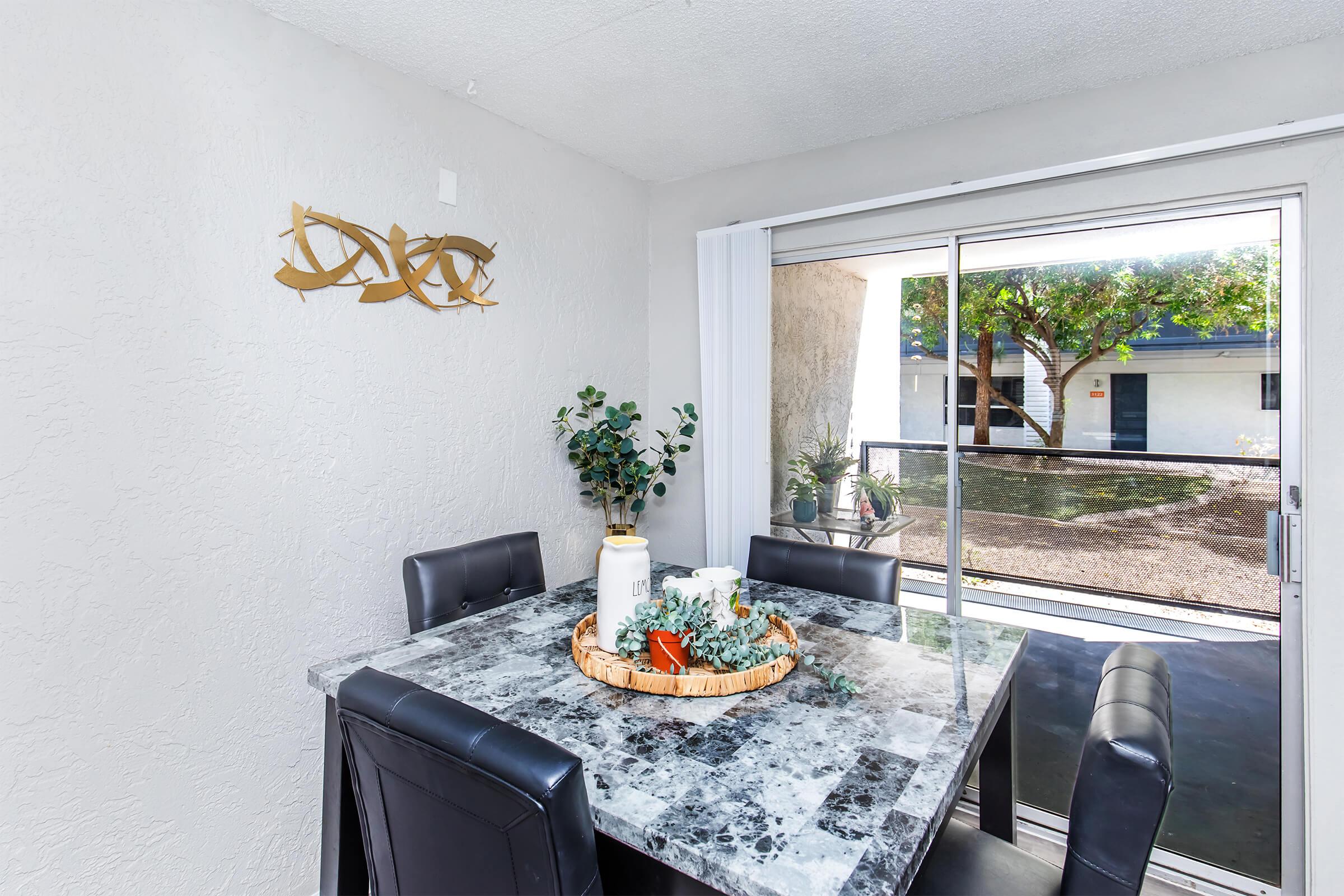A modern dining area featuring a marble table surrounded by black leather chairs. On the table, there is a decorative centerpiece with greenery. A sliding glass door leads to an outdoor view of trees and a patio. The walls are painted in a light color, and there is a decorative wall piece above the table.