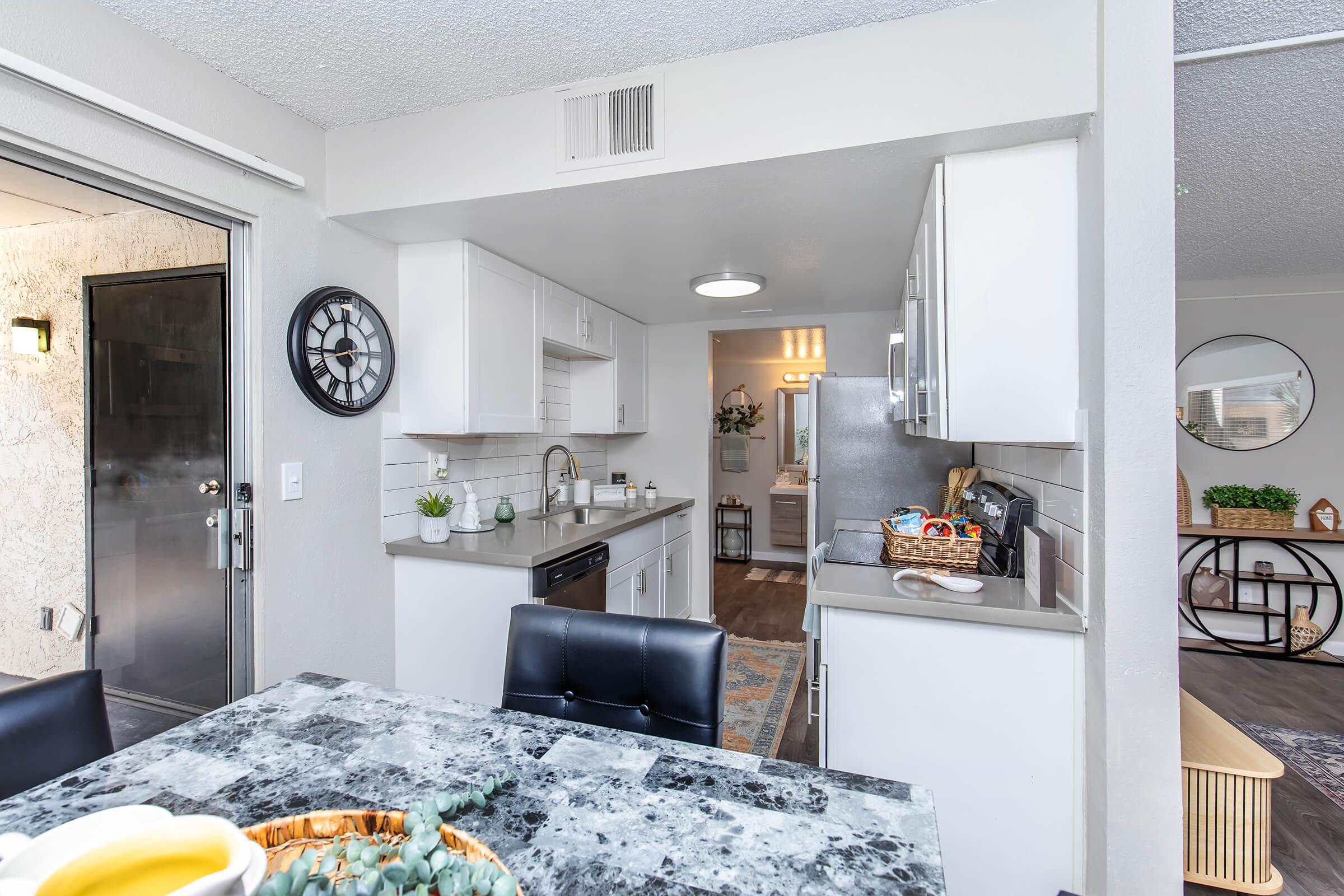Modern kitchen featuring white cabinetry, a gray countertop, and stainless steel appliances. A dining table with a decorative bowl sits nearby. The space is well-lit and open, leading to a hallway towards a bathroom and living area, with a door visible in the background.