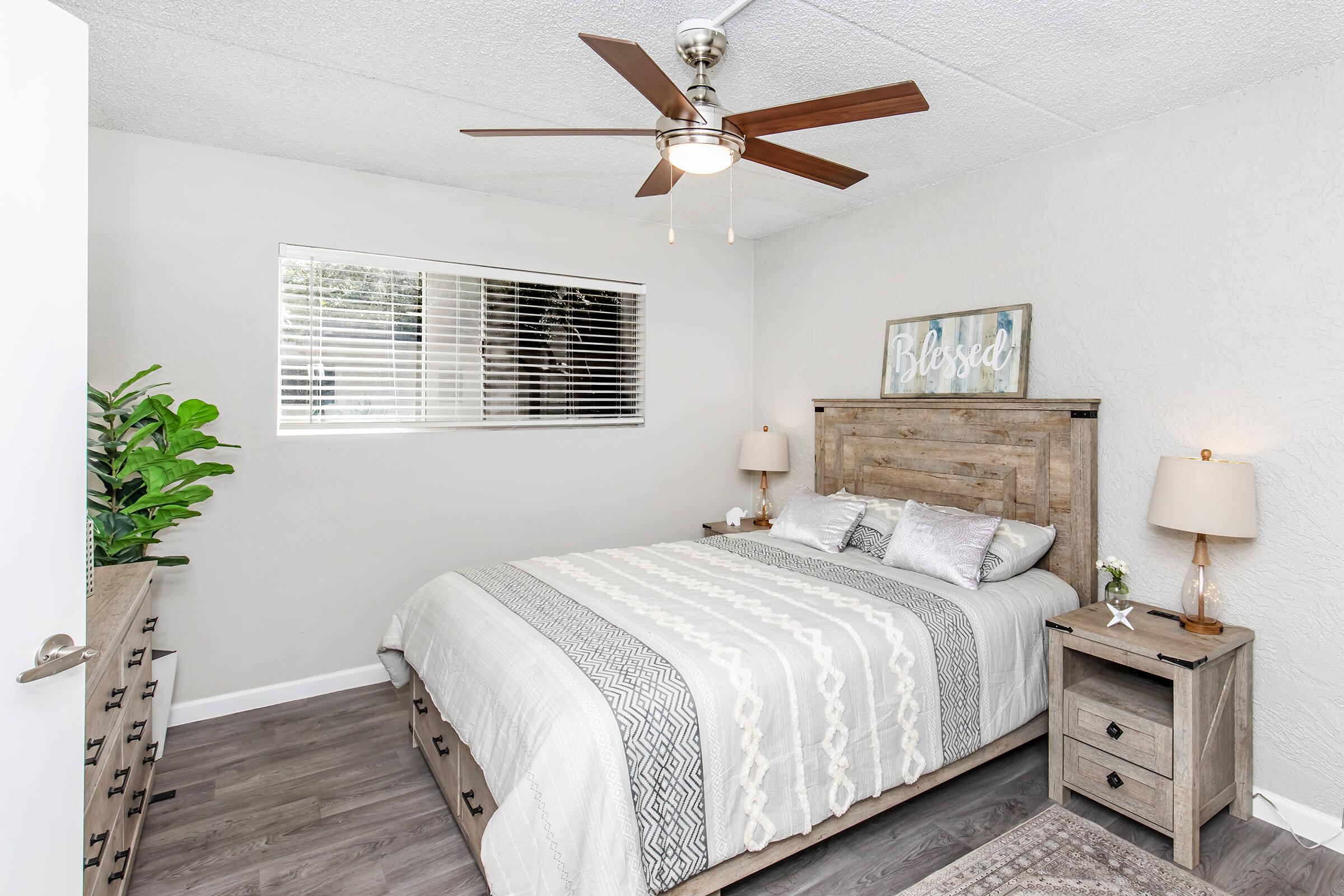 A cozy bedroom featuring a wooden bed frame with a light gray and white bedding set, two bedside lamps, a dresser, and a small plant. The room has a ceiling fan and a window with blinds, allowing natural light to enter. A decorative sign on the wall reads "Blessed."