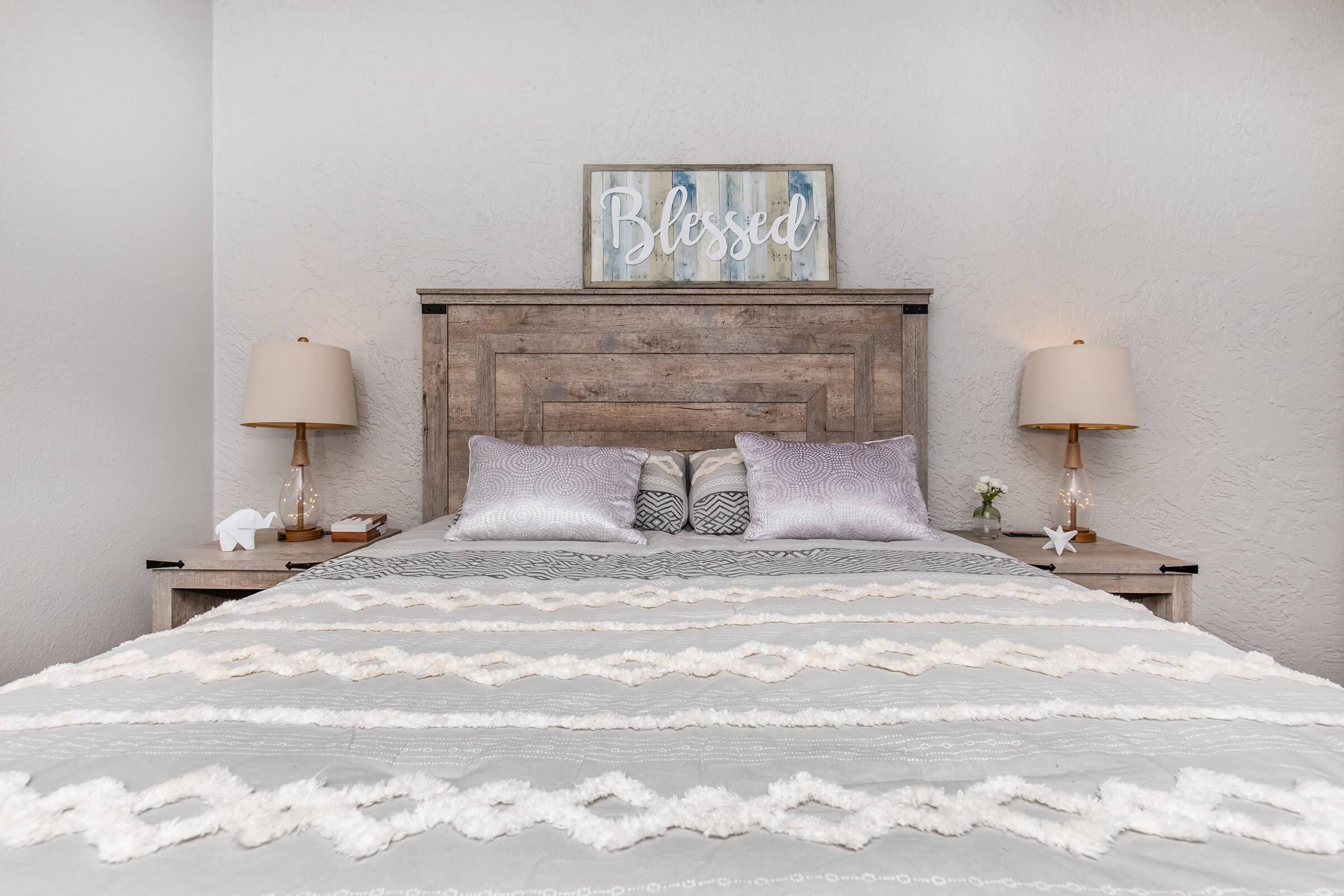 A cozy bedroom featuring a wooden headboard with a sign that reads "Blessed" above it. The bed is dressed in a light gray decorative blanket with textured patterns and fluffy pillows. Two bedside tables with lamps sit on either side, adding a warm, inviting ambiance to the space.