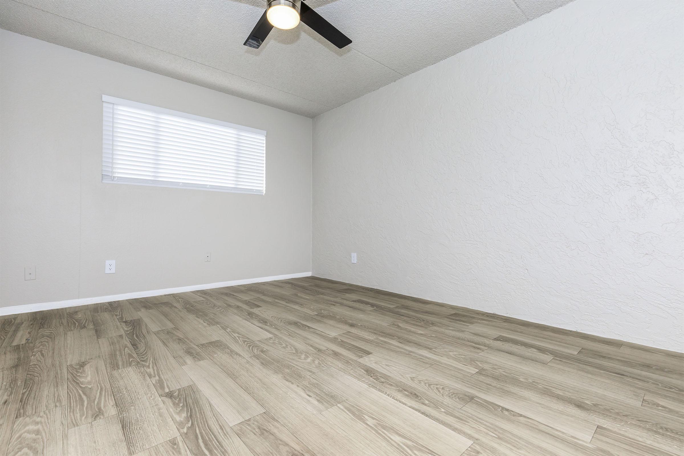 A spacious, empty room with light-colored walls and wood-like flooring. There is a small window with horizontal blinds on the left side, allowing natural light to enter. The ceiling features a modern ceiling fan, enhancing the room's air circulation. The overall ambiance is bright and minimalist.