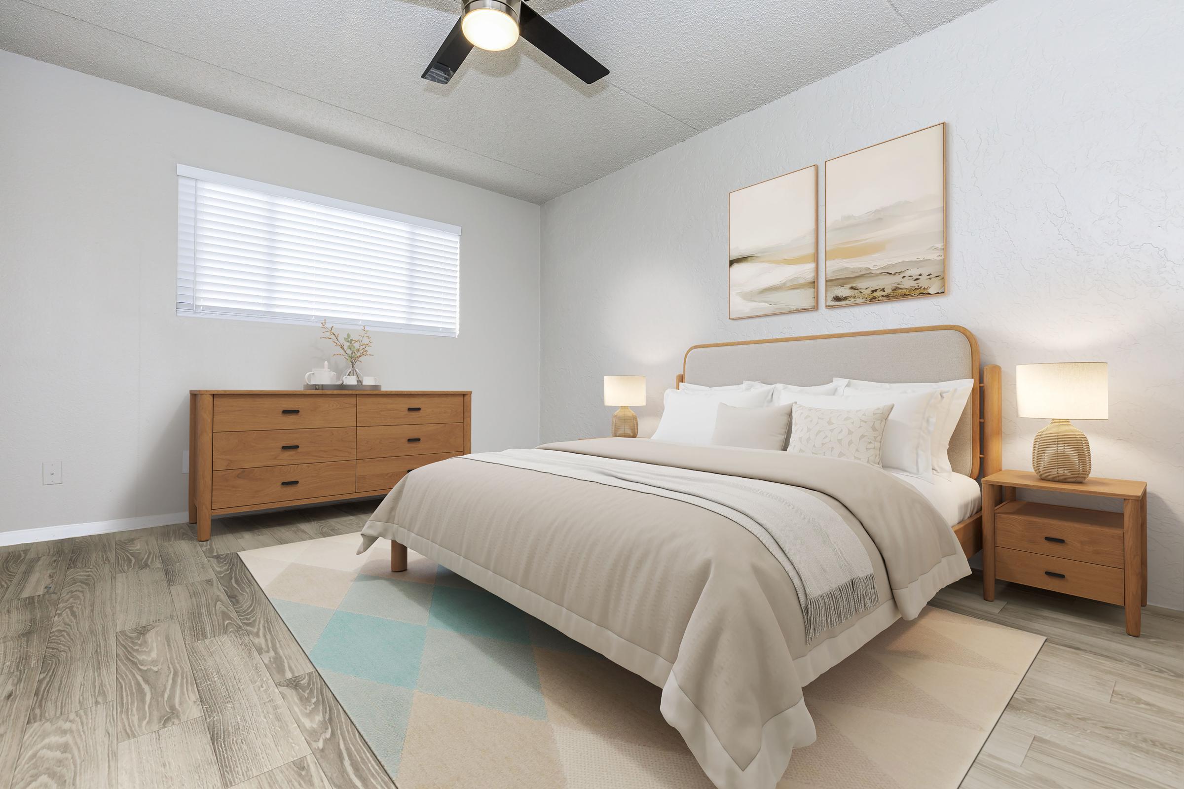 A modern bedroom featuring a large bed with neutral bedding, two decorative pillows, and a light-colored headboard. There are two bedside tables with lamps, a wooden dresser against the wall, and two framed art pieces above the bed. A window with blinds allows natural light to enter, and a soft, patterned rug lies on the floor.