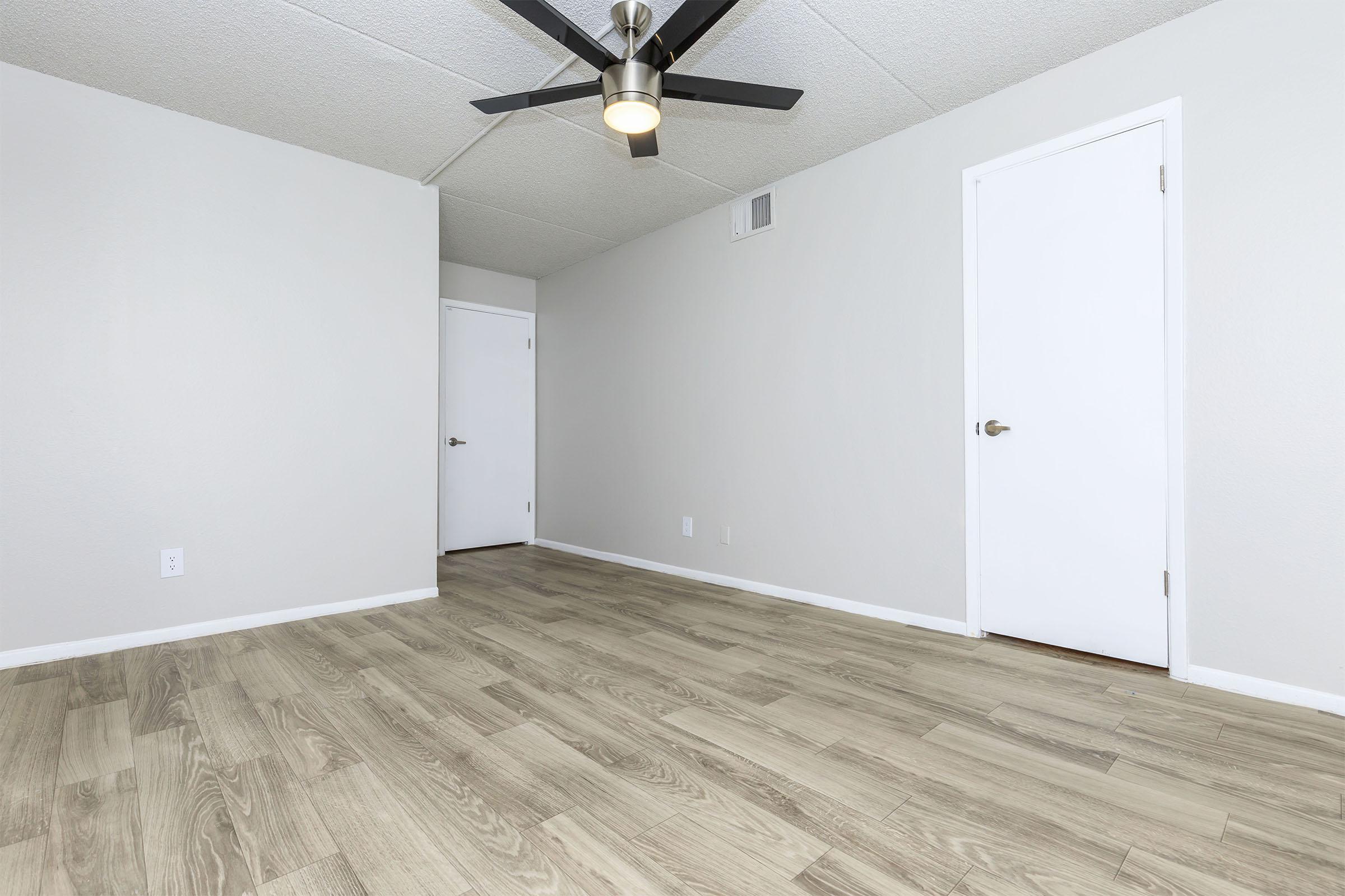 A spacious, empty room featuring light-colored walls and a wooden floor. A ceiling fan is mounted on the ceiling, and there are two doors visible on the right side. Natural light highlights the neutral decor, creating a bright and airy atmosphere.