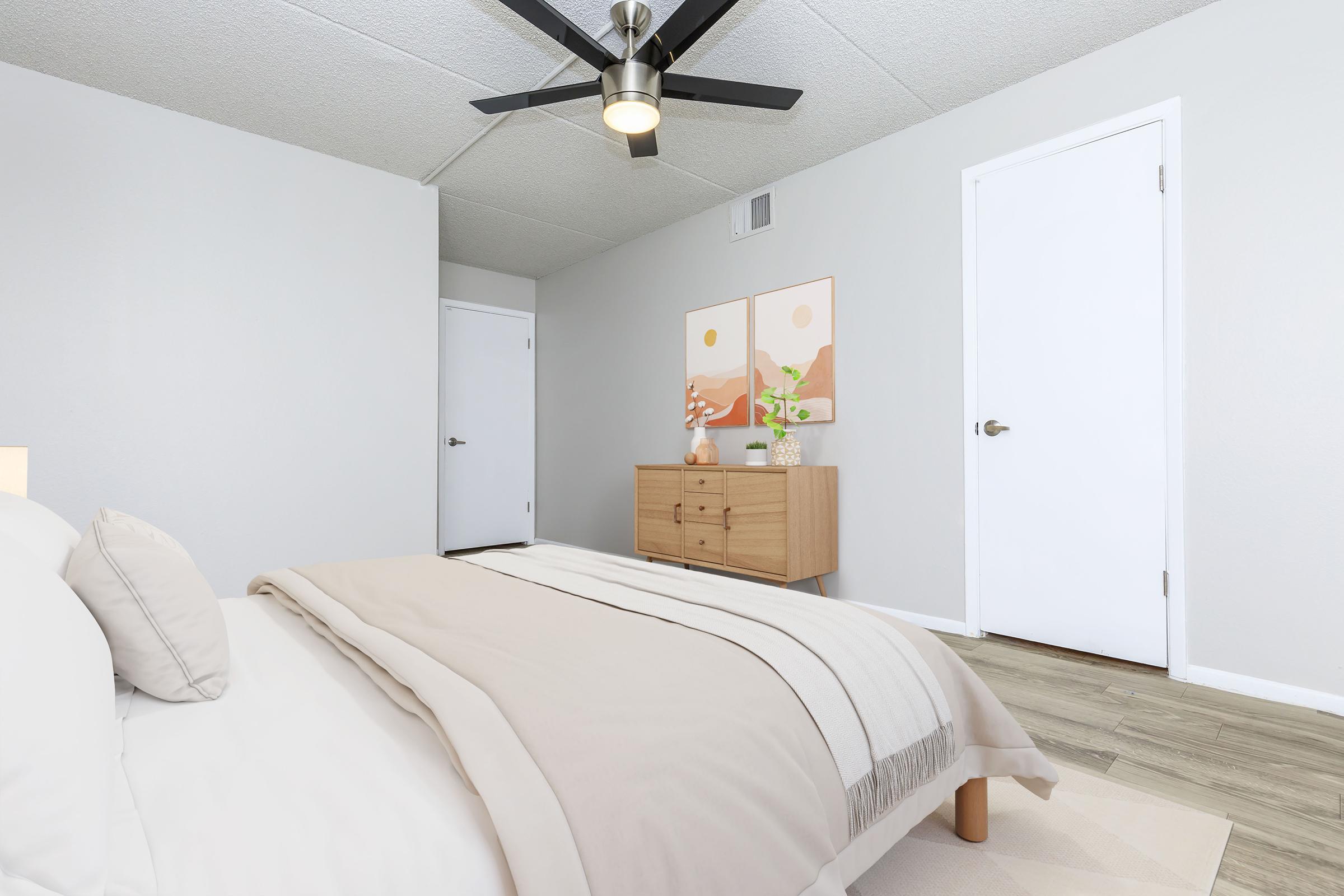 A cozy bedroom featuring a neatly made bed with beige linens, a light wood dresser, and two framed nature-themed artworks on the wall. There’s a ceiling fan above and a closed white door to the right, with a smooth, light-colored floor enhancing the calm atmosphere of the space.