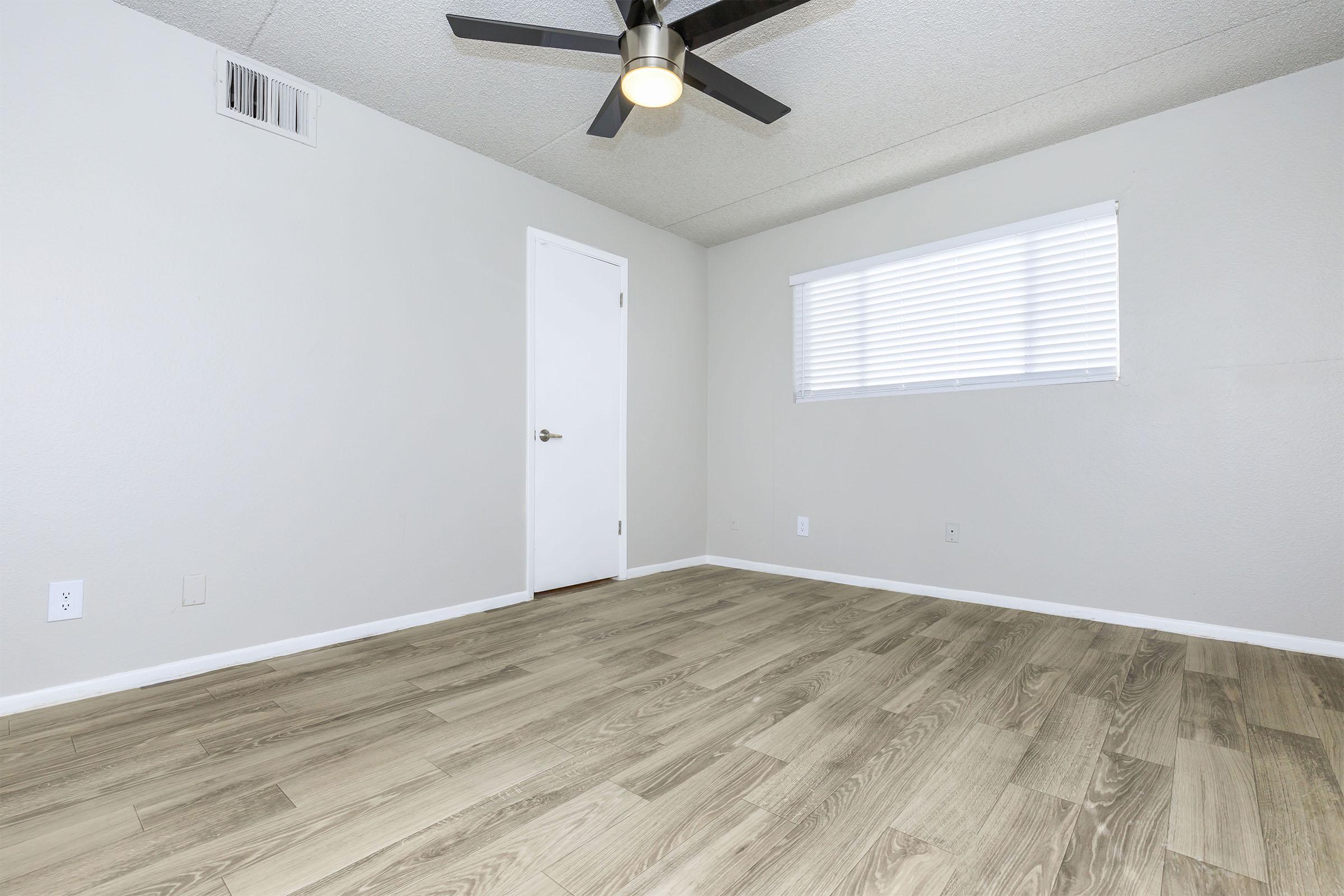 A spacious, empty room with light gray walls and a wooden floor. A ceiling fan is installed, and there's a window with white blinds allowing natural light. A closed white door is visible on one side of the room, enhancing the clean, minimalist aesthetic.