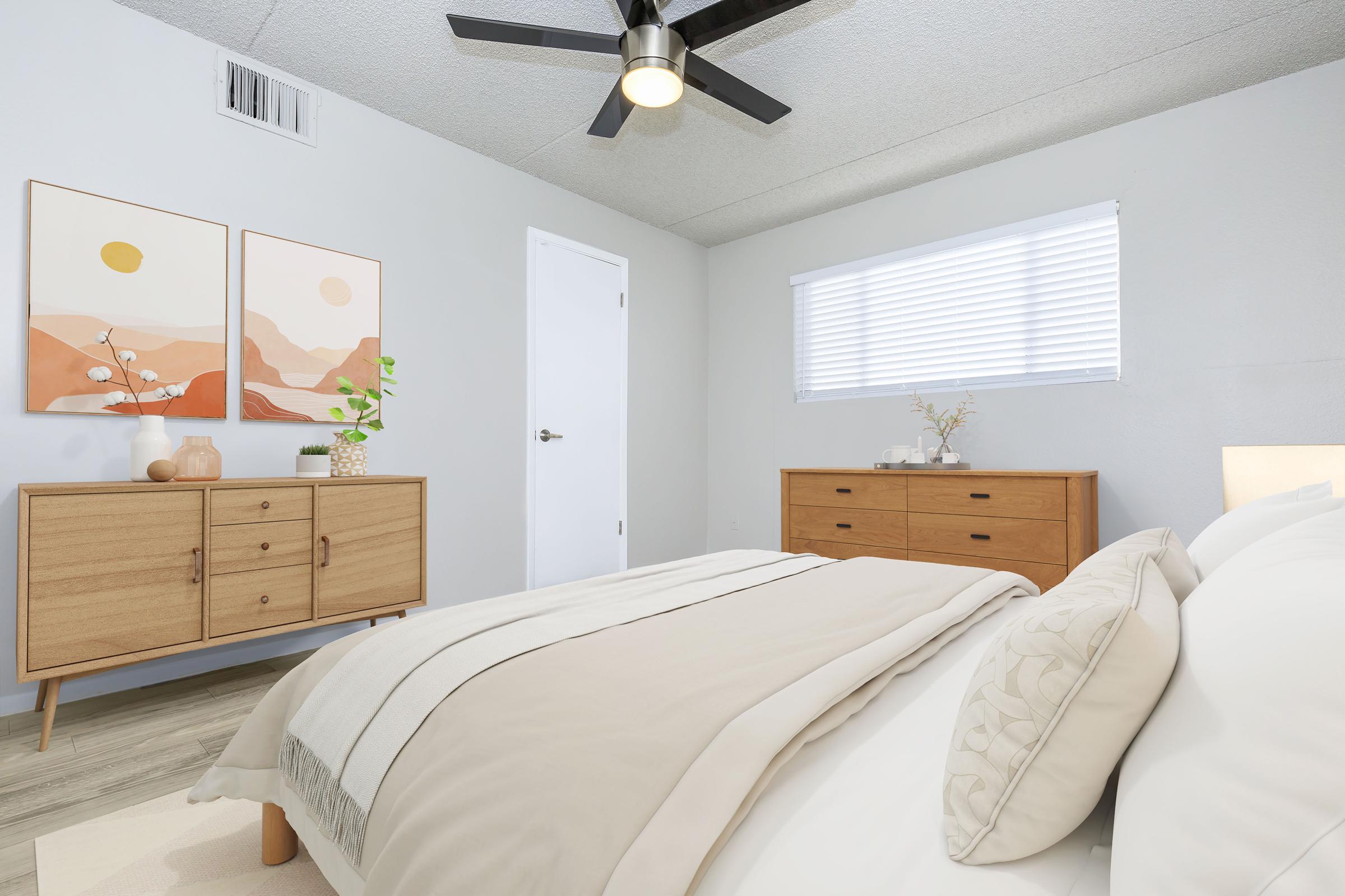 A cozy bedroom featuring a neutral color palette. It includes a bed with a light-colored blanket, wooden furniture, and decorative wall art depicting abstract landscapes. Natural light streams through a window with blinds, and a ceiling fan adds to the modern aesthetic. A plant and decorative items are placed on the furniture.